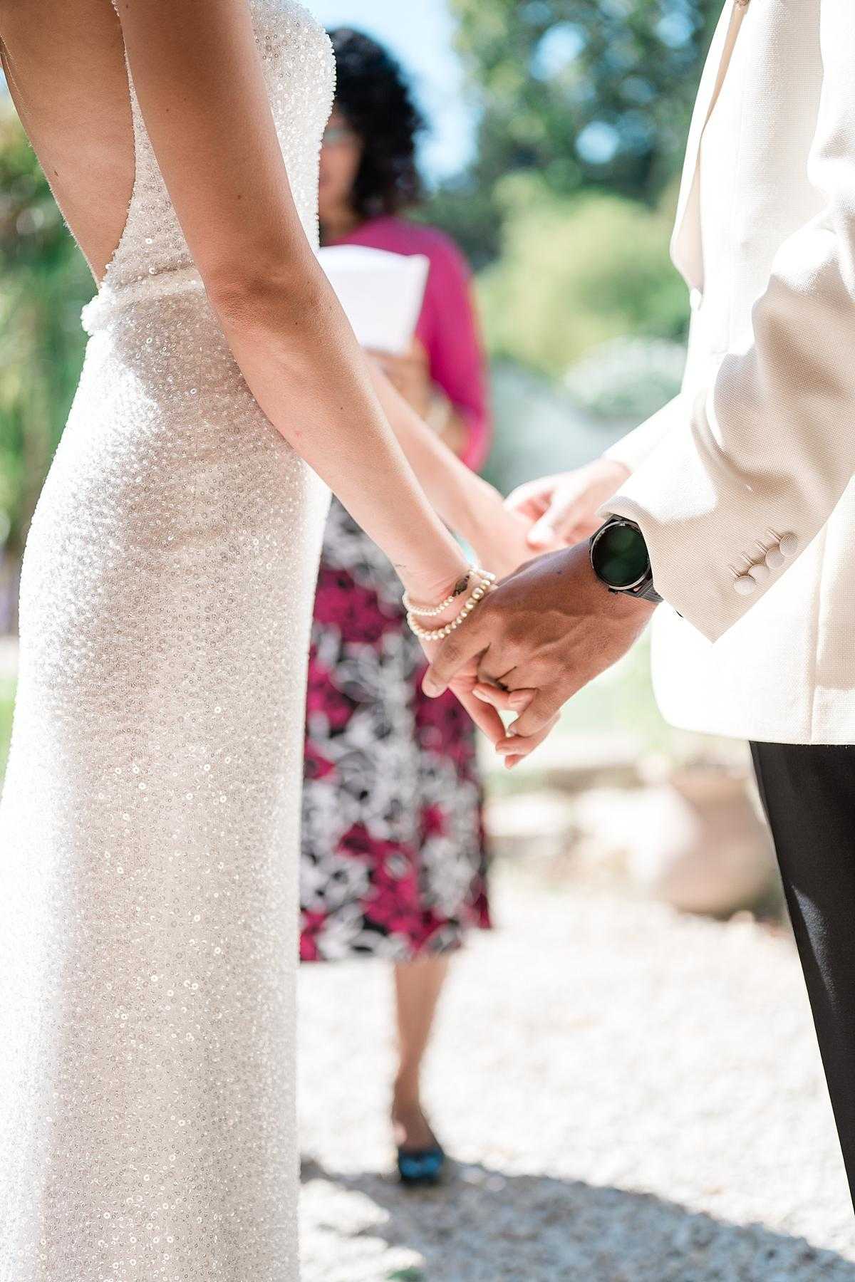 Close-up detail shot of a couple holding hands during an outdoor ceremony, with a celebrant or officiant visible and slightly out of focus in the background holding a white booklet and wearing a fuchsia jacket over a floral pink, white, and black skirt. The bride wears a fitted, all-over sequined ivory gown with a low open back, and her wrist is adorned with a pearl bracelet; the groom wears a cream or ivory blazer with dark trousers and a black smartwatch. The ceremony appears to take place in a garden or grounds setting in bright daylight.
