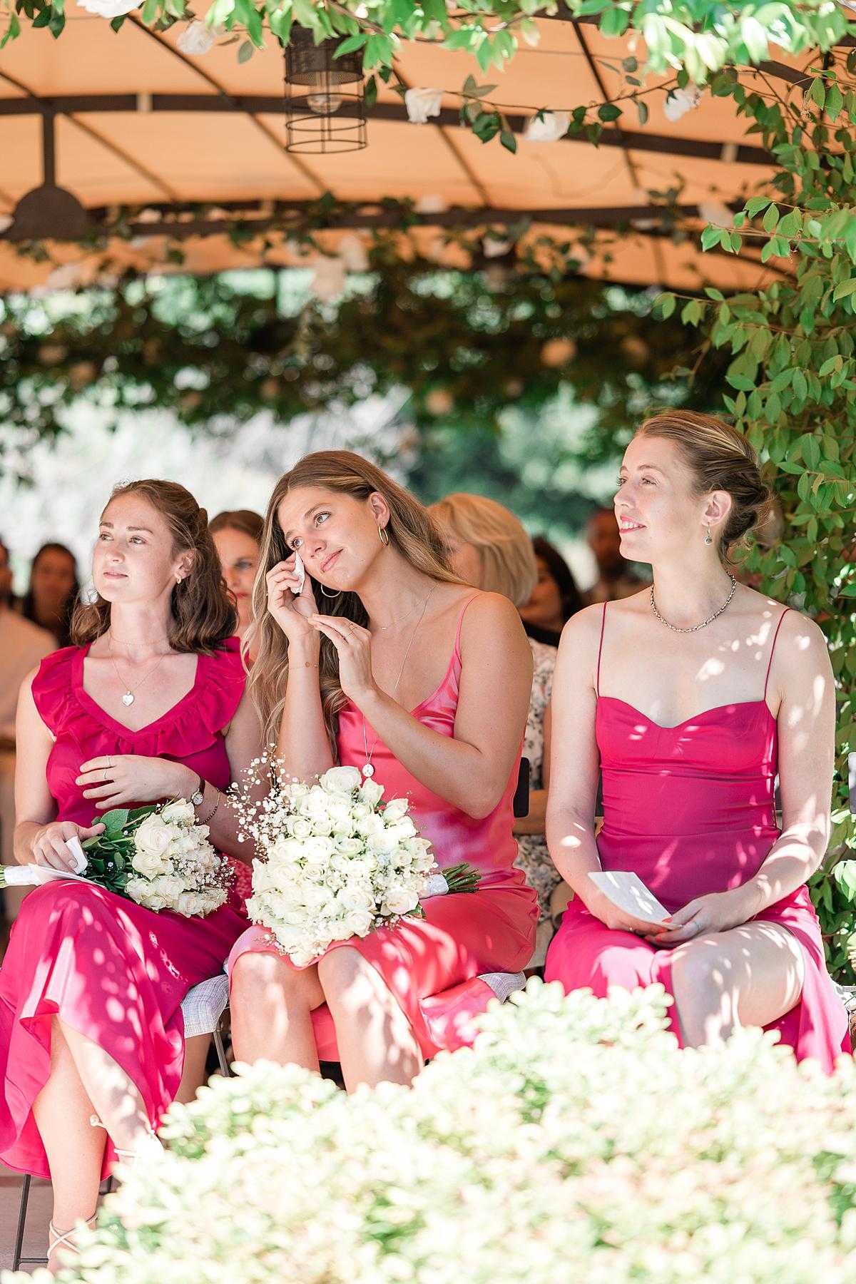 Three bridesmaids are seated during an outdoor wedding ceremony held under a cream-toned tent or pergola draped with trailing green foliage. All three women are wearing varying shades of hot pink and fuchsia dresses in different silhouettes — one with ruffled shoulders, one in a satin slip style, and one in a satin cowl-neck design — holding bouquets of white roses and baby's breath. The central bridesmaid is visibly emotional, wiping a tear from her eye, while the others watch the ceremony attentively. The foreground features white floral arrangements, and additional guests are visible seated in the background. The image is a mid-range portrait shot with soft dappled sunlight filtering through the foliage overhead.
