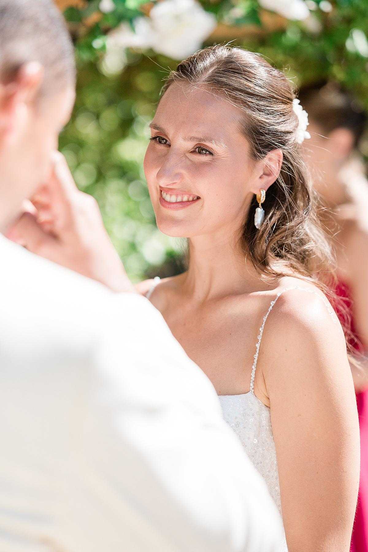 A close-up portrait of a bride during an outdoor ceremony, smiling warmly at the groom whose back is blurred in the foreground. The bride wears a white spaghetti-strap dress with subtle embellishment, gold hoop earrings with a pale blue stone drop, and a small white flower pinned in her loosely styled half-up hair. In the background, a white floral arch and foliage are softly out of focus, along with what appears to be a guest in a red outfit. The shot is taken over the groom's shoulder, creating an intimate over-the-shoulder composition with shallow depth of field.
