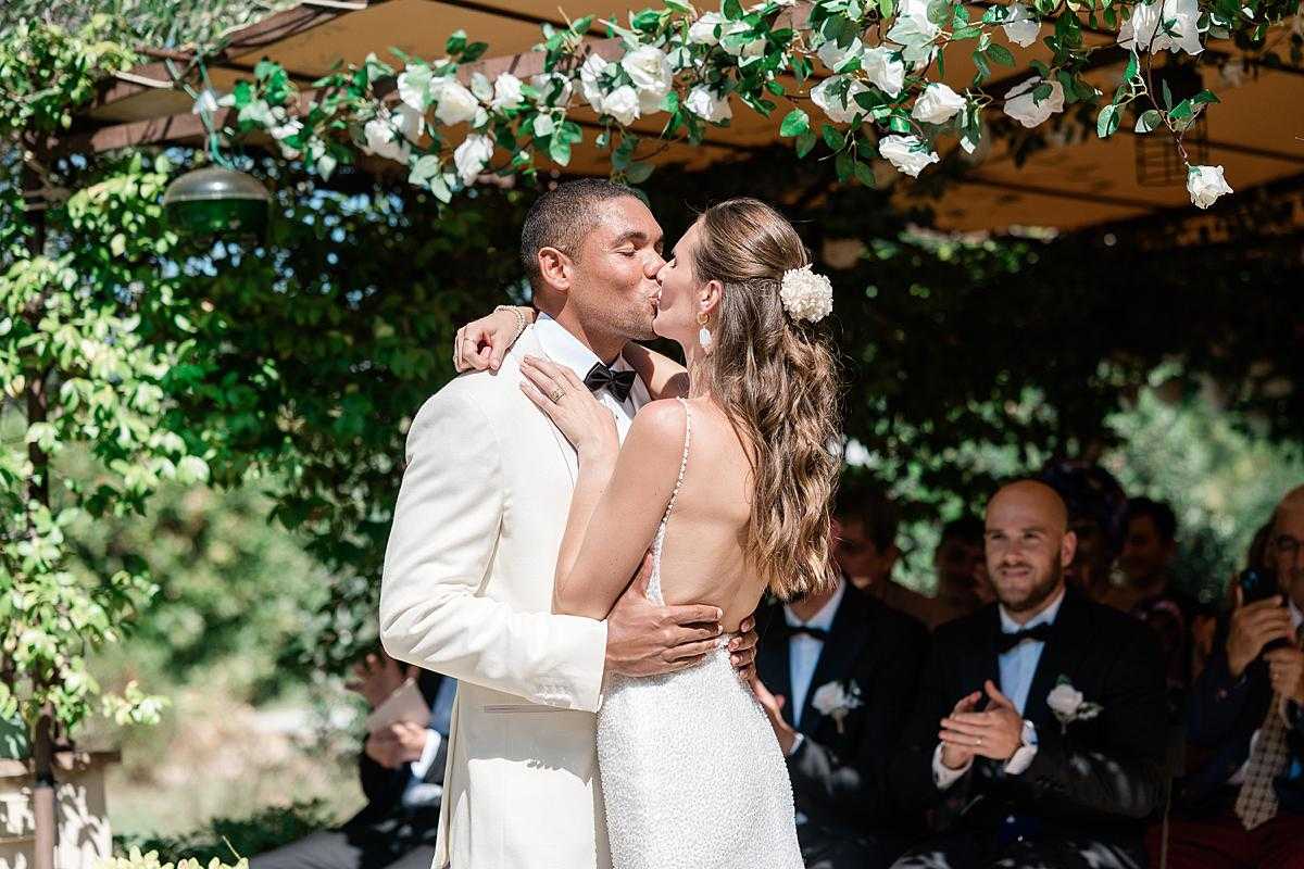 The couple shares their first kiss during an outdoor ceremony beneath a pergola or covered structure draped with white roses and green foliage. The groom wears an ivory dinner jacket with a black bow tie, while the bride wears a fitted, low-back white beaded or textured gown with spaghetti straps and has a white floral hair accessory — likely a small white chrysanthemum or ranunculus — pinned into her loose wavy hair. Seated guests, including men in navy suits with white boutonnieres, are visible in the background applauding, with one guest photographing the moment on a phone. The shot is a medium portrait-style frame capturing the couple from the waist up, with the ceremony arch of white roses and greenery framing the top of the image.
