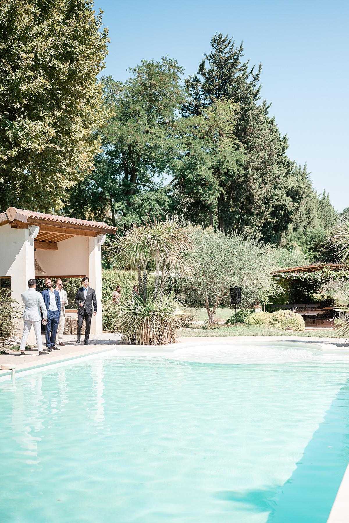 Outdoor cocktail hour scene at a Provençal-style property, photographed as a wide shot from poolside level. A large rectangular swimming pool with blue-green water occupies the foreground, while three male guests in suits — one in a light grey linen suit, one in navy blue, and one in dark charcoal — stand conversing beside a pool house with terracotta roof tiles and white render walls. A small group of additional guests is visible in the background near a pergola structure on the right. The setting features a classic southern French outdoor leisure space with a relaxed, sunny daytime atmosphere. Potential venue feature image.