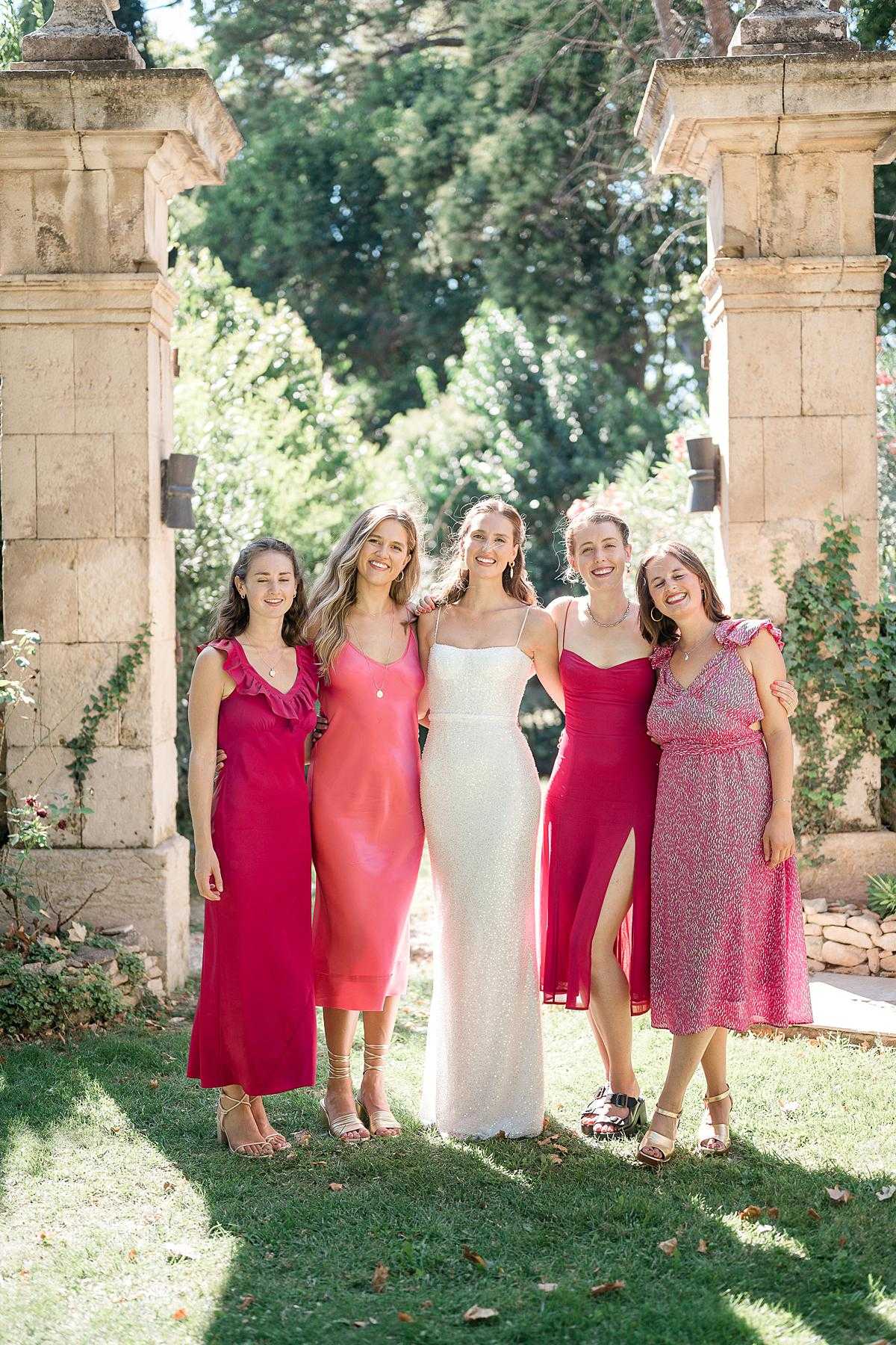 A bridal party portrait taken outdoors at a French chateau or domaine, with the bride and four bridesmaids posing together in front of a stone pillar gateway with ivy climbing the columns. The bride wears a fitted, spaghetti-strap white sequined gown, while the bridesmaids wear mismatched dresses in a coordinated pink-to-red palette: a hot pink ruffled midi, a coral satin slip dress, a crimson satin midi with a side slit, and a pink floral print midi with ruffled sleeves. All five women are smiling and have arms around each other in a relaxed, candid pose. The setting is a sun-drenched garden with a classic stone archway, giving the image a classic French estate feel. Full-length portrait shot in natural daylight.