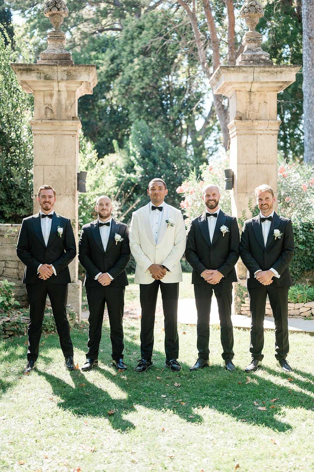 A groomsmen portrait taken outdoors in the grounds of what appears to be a French chateau or estate, with two large ornamental stone pillars topped with carved urns framing the group. The groom stands at center wearing an ivory dinner jacket with black trousers and a black bow tie, while his four groomsmen wear matching all-black tuxedos with black bow ties. All five men wear small white floral boutonnieres and stand with hands clasped in front, smiling toward the camera. The composition is a classic full-length portrait shot in bright natural daylight on a manicured lawn.