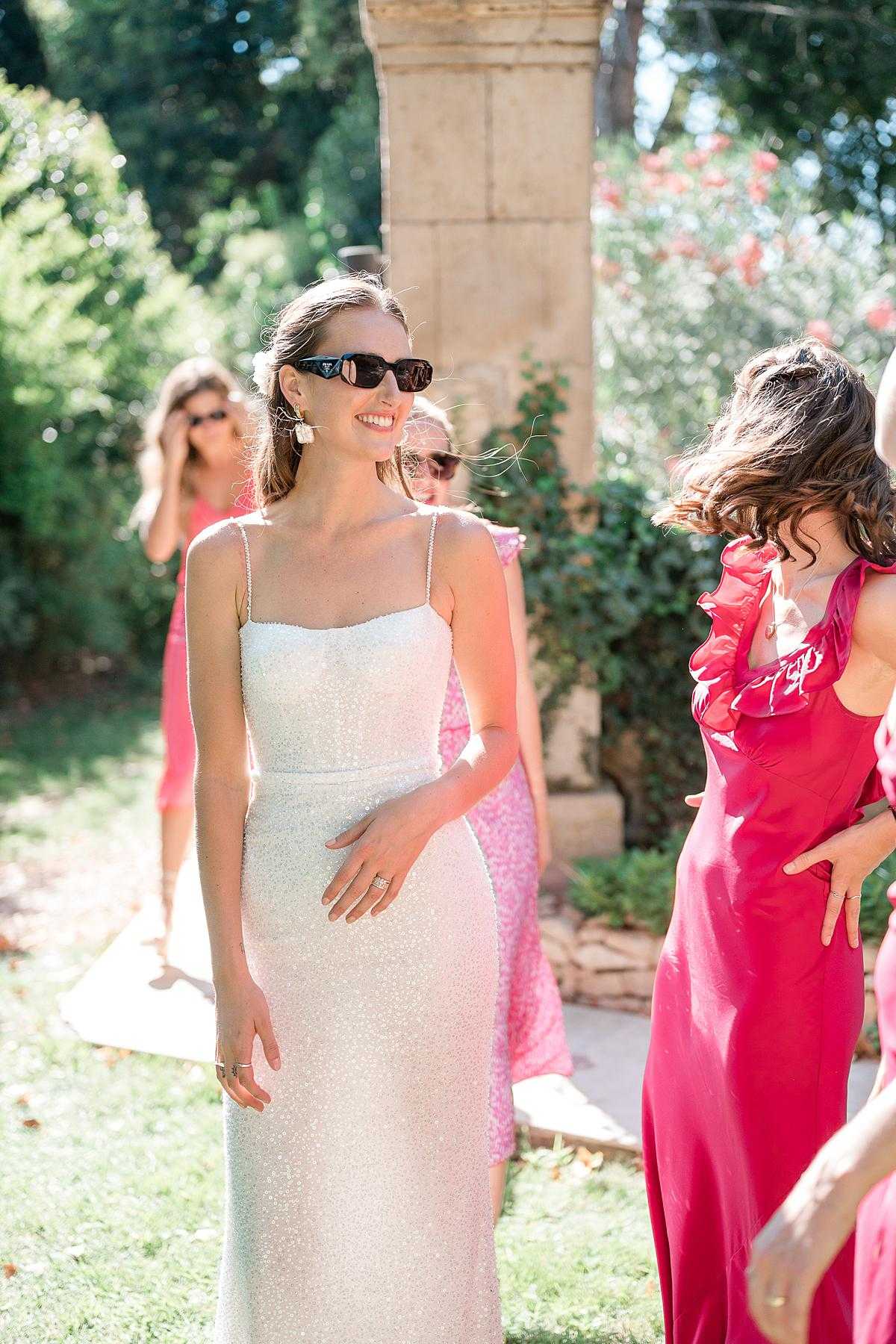 A bride stands outdoors in a garden setting, smiling and wearing a slim-fit ivory sequined gown with thin spaghetti straps, paired with black rectangular sunglasses and statement drop earrings. She is surrounded by female guests dressed in shades of hot pink and fuchsia, including one woman in a bold red-pink satin gown with ruffle shoulder detail visible to the right. The setting appears to be the grounds of a French estate or chateau, with a stone architectural column visible in the background. The shot is a mid-length portrait taken in bright natural sunlight, with the background softly blurred.