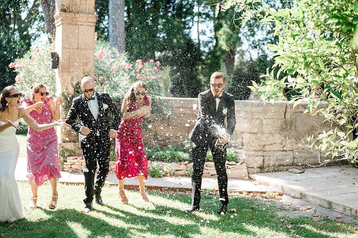 A candid outdoor reception moment in a garden or courtyard setting, where a group of five guests are reacting with excitement as a champagne bottle is being sabered or shaken, sending a dramatic spray of liquid and bubbles through the air. The guests include two women in hot pink and red midi dresses and a woman in a white dress, alongside two men in black tuxedos with bow ties, all wearing sunglasses. The setting features a stone wall, paved terrace, and manicured lawn in bright midday sunlight. Wide shot with strong action energy and high contrast from the direct sun.