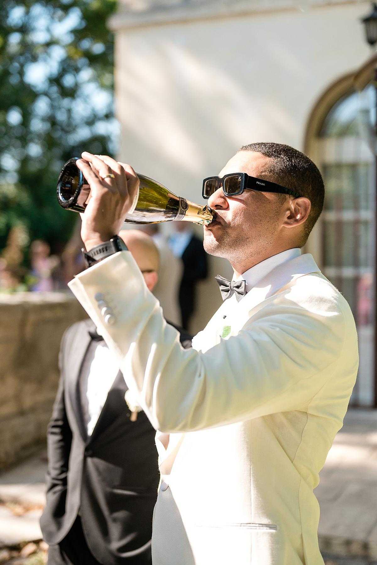 A candid outdoor shot capturing a groom drinking directly from a champagne bottle during what appears to be a cocktail hour or post-ceremony celebration. The groom is dressed in a cream ivory dinner jacket with a grey bow tie, a white boutonniere, and is wearing black Prada sunglasses, giving the moment a playful, stylish tone. In the background, another man in a dark suit is visible along with blurred guests, set against the arched facade of what appears to be a French chateau or manor house. The image is a mid-range portrait shot taken in bright natural sunlight.