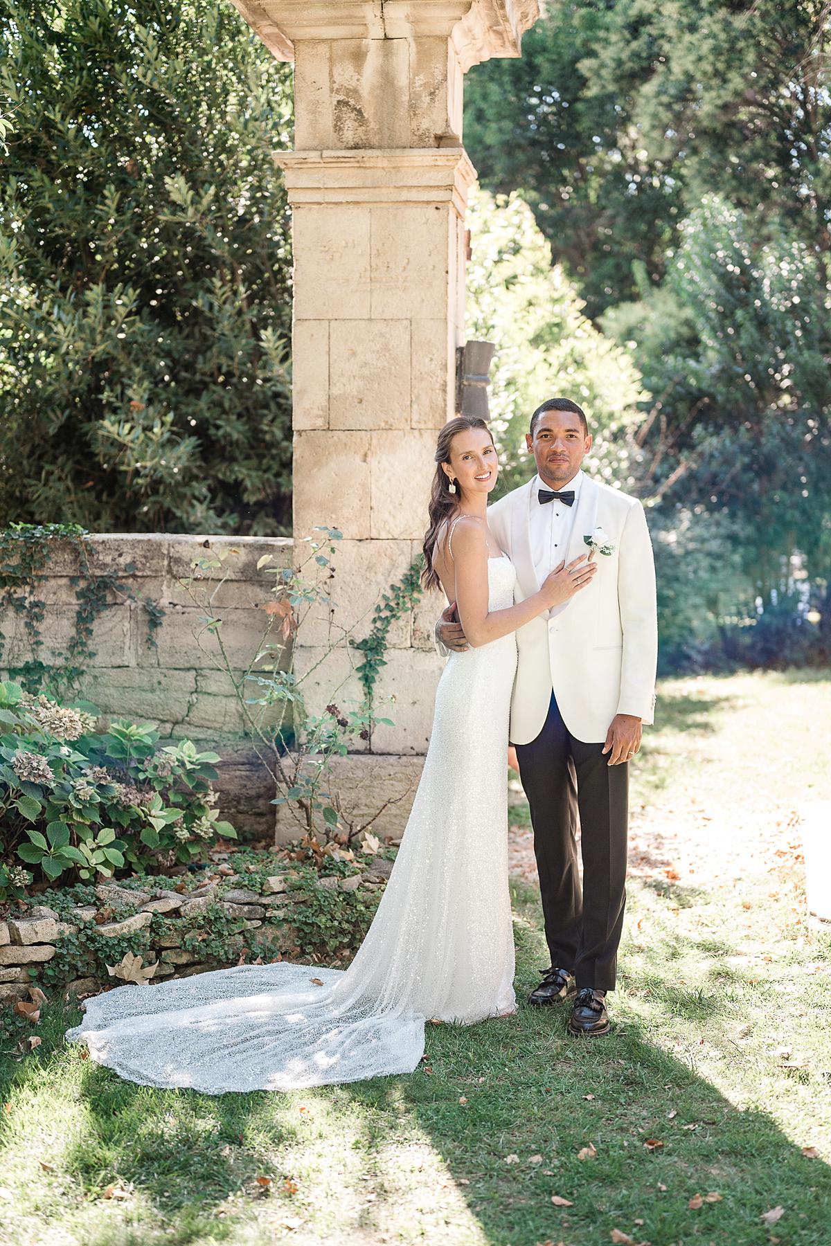 A couple portrait taken outdoors in the grounds of what appears to be a French chateau or estate, with a large stone column and low stone balustrade visible behind them. The bride wears a fitted, full-length ivory sequined or beaded gown with a long trailing train and thin spaghetti straps, paired with drop earrings and her dark hair loosely styled over one shoulder. The groom wears a cream dinner jacket with a black bow tie, dark navy or black trousers, black dress shoes, and a small white boutonniere. The bride rests her hand on the groom's chest as he stands slightly behind her, with natural dappled sunlight casting soft shadows across the scene. The overall styling is classic and formal. Full-length couple portrait shot.