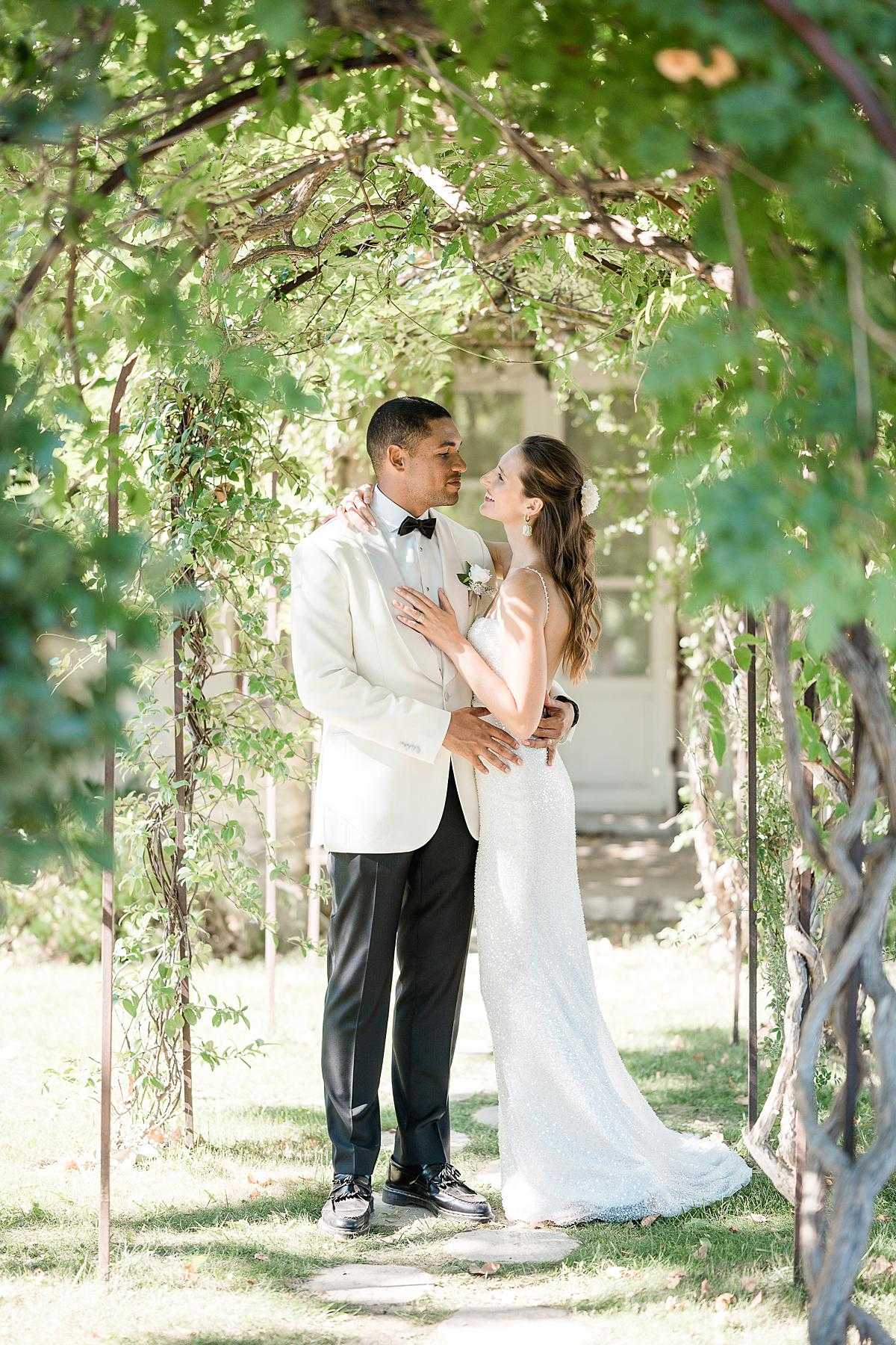 A couple portrait taken outdoors in a garden setting, with the bride and groom standing beneath a vine-covered metal arch. The groom wears an ivory dinner jacket with black trousers, a black bow tie, and a white boutonniere, while the bride wears a fitted, sleeveless white sequined gown with a small white floral hair accessory and her hair styled in loose waves. The two are facing each other closely, with the bride's hand resting on the groom's chest and his hands at her waist. A white building is partially visible in the background through the archway. The shot is a full-length portrait with soft natural light filtered through the foliage overhead, giving the image a bright, airy quality.