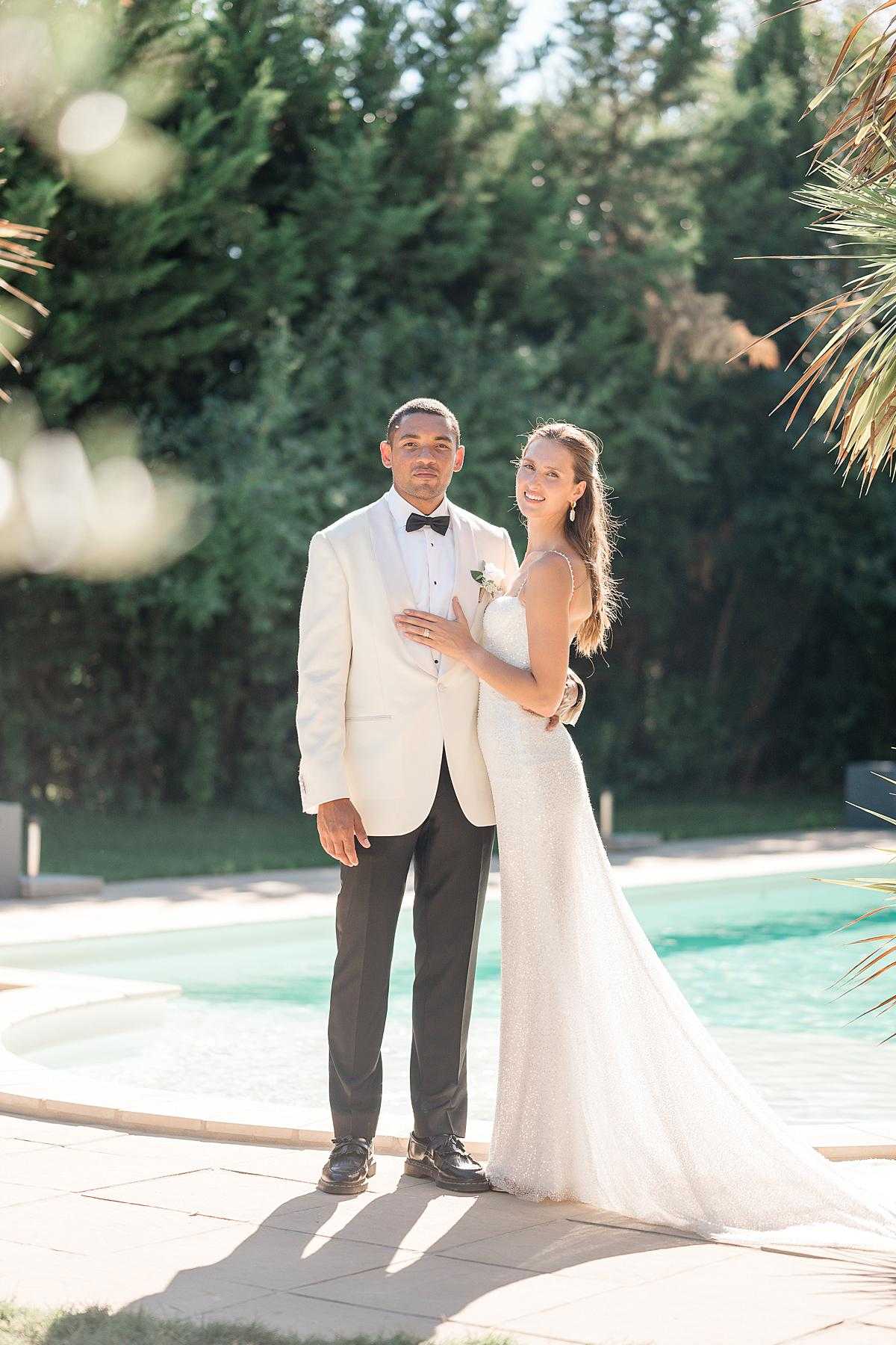 A couple portrait taken outdoors beside a swimming pool, likely during a post-ceremony portrait session. The groom wears a cream/ivory dinner jacket with black lapels, a black bow tie, black trousers, and black dress shoes, with a small white floral boutonnière. The bride stands slightly behind him with her hand resting on his chest, wearing a form-fitting, full-length sparkle or sequined ivory gown with thin spaghetti straps and a short train; her hair is worn down. The bride smiles toward the camera while the groom faces forward with a more neutral expression. The setting is an outdoor pool area with stone paving, framed by palm fronds in the foreground and tall cypress or conifer trees in the background, in warm afternoon sunlight. The overall styling is modern classic. The shot is a full-length couple portrait with soft bokeh lens flare visible in the upper left corner.