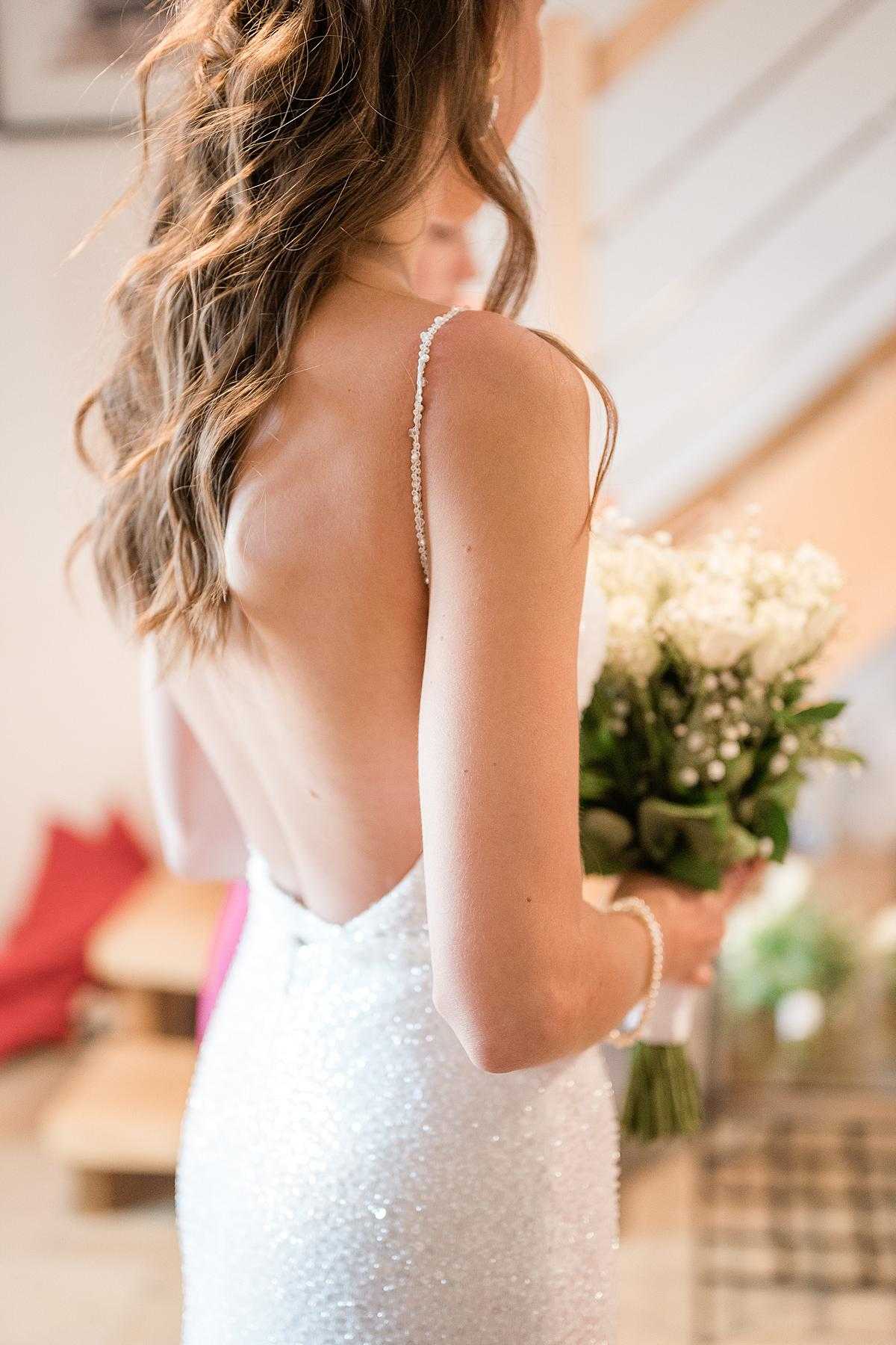 A close-up portrait of a bride shot from behind and slightly to the side, indoors in what appears to be a getting-ready space with soft natural light. She wears a white sequined fitted gown with a deep open back and a thin crystal-embellished spaghetti strap, and a delicate pearl bracelet on her wrist. Her dark brown wavy hair falls loosely over one shoulder. She holds a compact bouquet of white tulips, lily of the valley, and dark green foliage with a green ribbon-wrapped stem, looking downward with her face partially turned away from the camera.