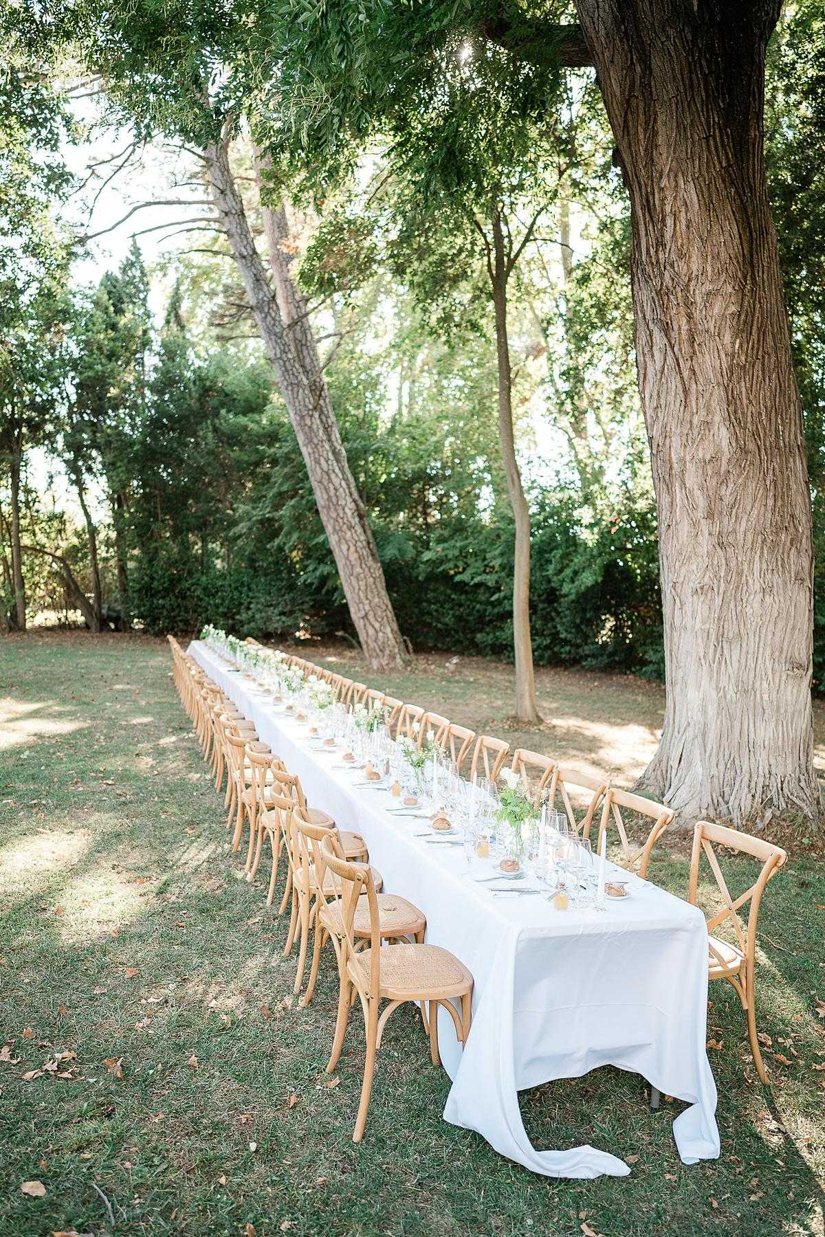 An outdoor wedding reception table setup photographed in a wide shot from a slightly elevated angle. A single long rectangular banquet table, draped in a white linen tablecloth, is set on a garden lawn and lined on both sides with natural wood cross-back chairs with woven rattan seats. The table is set for approximately 30 guests and features glassware, grey linen napkins, small bread rolls, and a low centerpiece runner of delicate white flowers and greenery in clear bud vases running the length of the table. The overall decor palette is white, natural wood, and soft green, with a clean, classic-rustic styling.