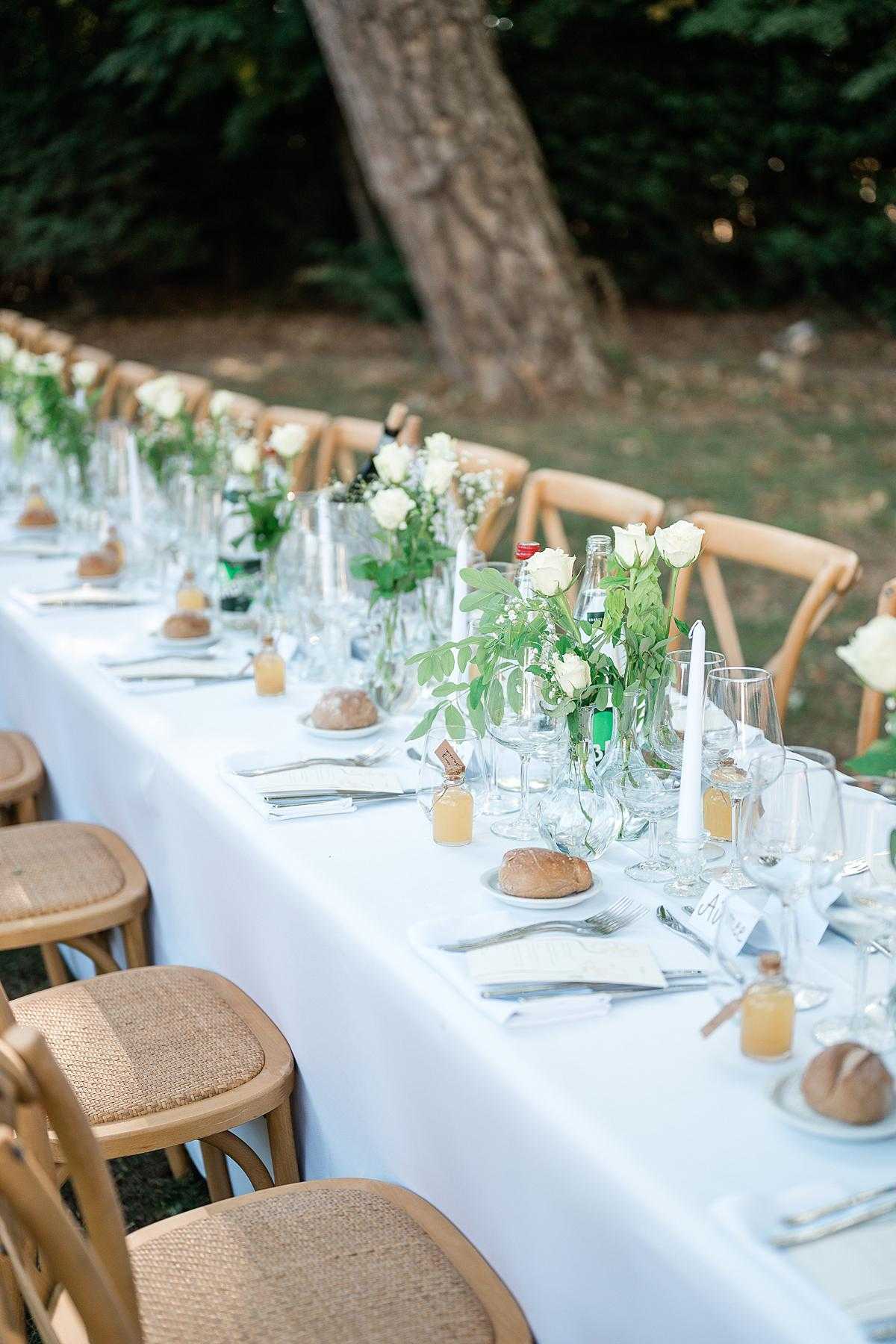 A detail shot of an outdoor wedding reception long table set for a seated dinner, photographed from a low angle along the length of the table. The table is dressed with a white linen cloth and lined with natural wood cross-back chairs with woven rush seats. Centerpieces consist of clear glass vases holding white roses and loose green foliage, interspersed with slim white taper candles, water bottles, and multiple clear wine and water glasses per place setting. Each place setting includes a small bread roll on a white plate, silverware wrapped in a white napkin, a printed menu card, and a small honey jar favor with a cork stopper. The styling is relaxed and natural, with a green-and-white color palette and a rustic-meets-classic aesthetic.