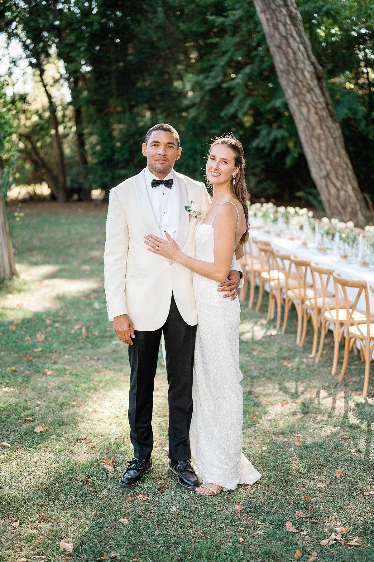 A couple portrait taken outdoors in a garden setting, with the bride and groom posing together facing the camera. The groom wears a cream/ivory dinner jacket with black lapels, a black bow tie, black trousers, and a small white floral boutonniere with greenery; the bride wears a fitted, spaghetti-strap ivory sequined gown and has her hair half-up. In the background, a long outdoor reception table is set with a white linen runner, tall glassware, and low white floral centerpieces with greenery, lined on both sides with natural wood cross-back chairs. The overall styling is classic and modern with a neutral white and green palette. The image is a medium full-length portrait shot with natural daylight.