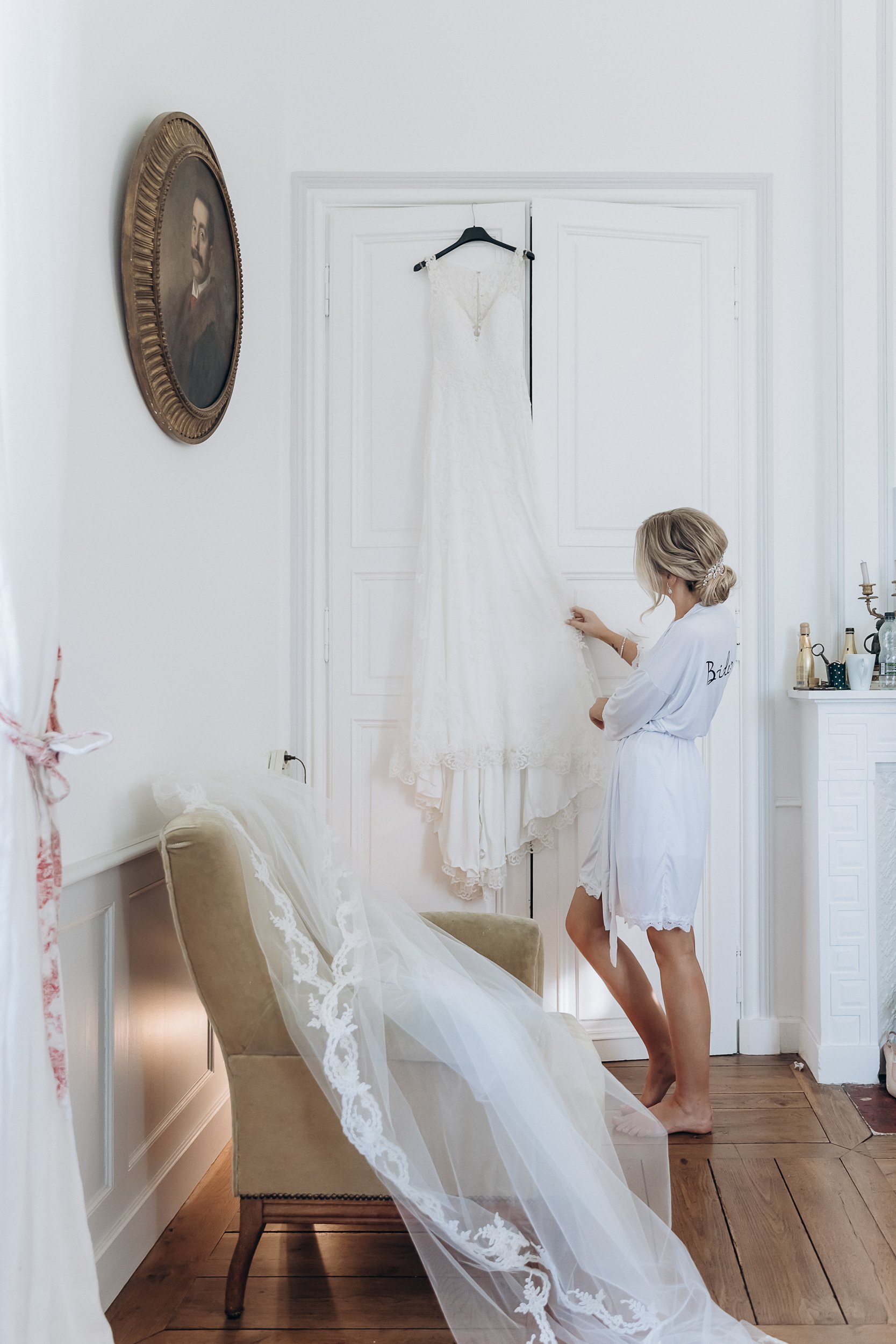 A getting-ready scene captured indoors in a white-walled room with classic French interior details including panelled doors, a white fireplace mantel, and wide-plank hardwood floors. The bride, wearing a white satin robe embroidered with 'Bride' on the sleeve and her blonde hair pinned up with a decorative hair piece, stands barefoot examining her ivory lace wedding gown, which hangs from a black hanger on the door. In the foreground, a long cathedral-length veil with wide lace appliqué trim is draped over an antique sage velvet armchair. A gold-framed oval portrait painting hangs on the wall to the left, adding to the classic French chateau aesthetic of the room. The shot is a full-length wide portrait taken from a low, slightly distanced angle, incorporating both the dress and the veil as separate focal elements.