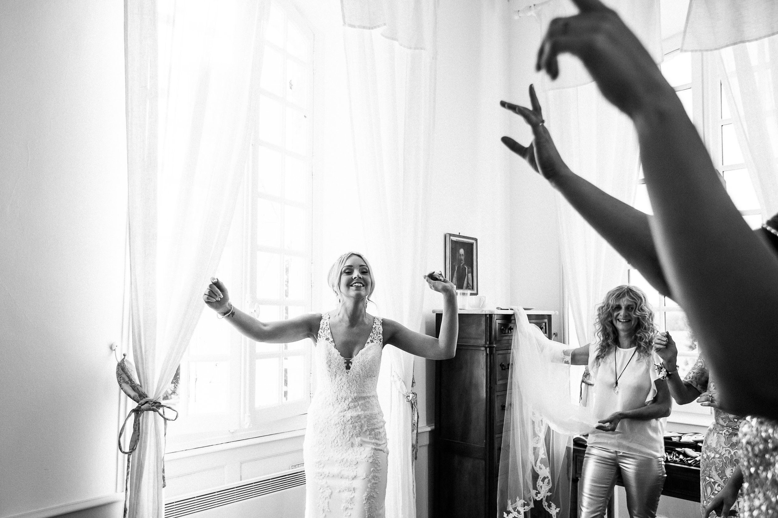 A black-and-white getting-ready scene shot indoors, likely in a chateau or manor room, with tall sheer curtained windows providing bright backlighting and high contrast. The bride stands center frame with arms outstretched and a wide smile, wearing a fitted lace wedding dress with a deep V-neckline and lace appliqué detailing; two women assist in placing or adjusting her veil, with one woman's raised arms and hands in sharp foreground silhouette creating a dynamic compositional frame. A third woman with curly hair, dressed in a light top and metallic trousers, holds the edge of the lace-trimmed veil to the bride's right. The room features white walls, a dark wood antique cabinet, and a framed portrait on the wall, suggesting a classic French interior setting. The image is a wide-angle, slightly low-angle candid shot with strong tonal contrast between the bright window light and the shadowed foreground.