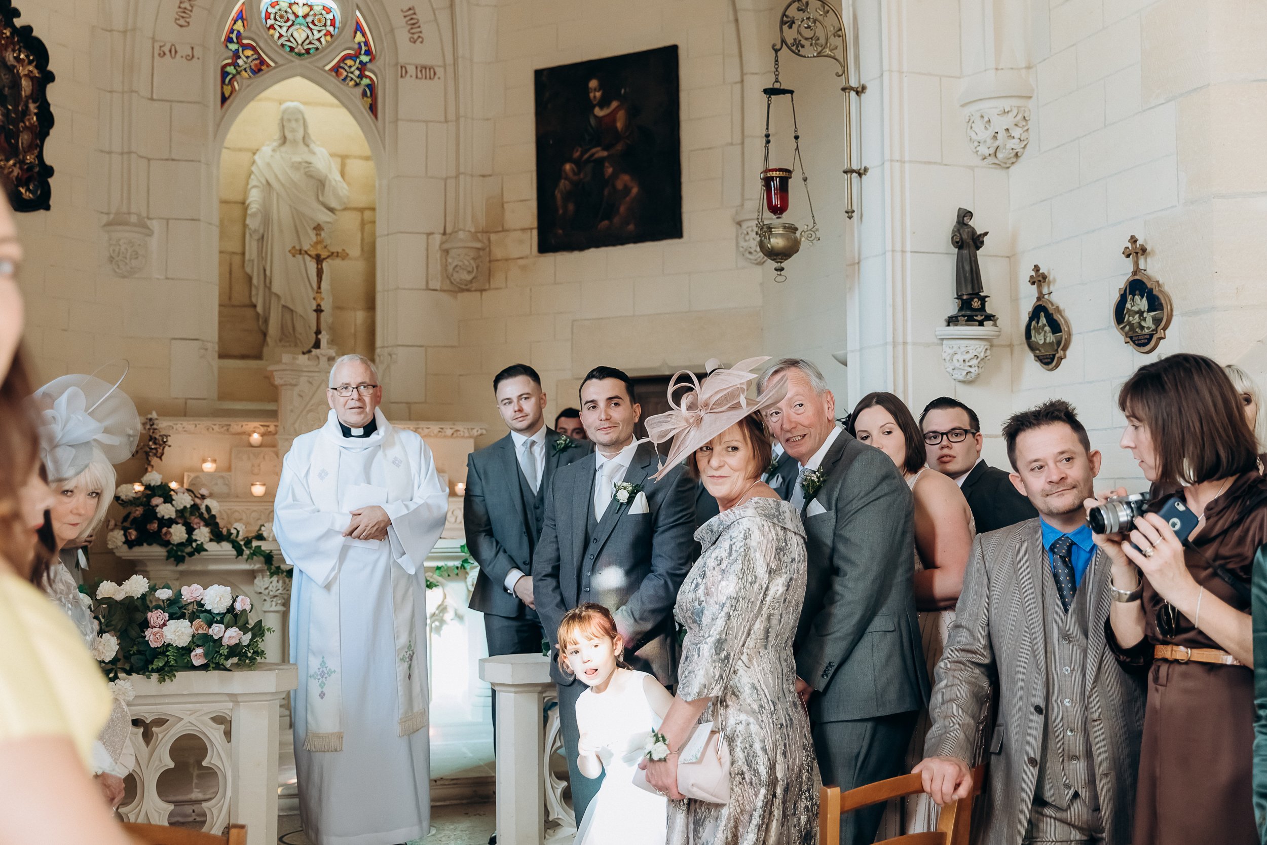 A wedding ceremony is taking place inside a French Catholic chapel with white limestone walls, Gothic arched alcoves, stained glass windows, religious statues, oil paintings, and candles lit at the altar. A priest in white vestments stands at the altar, while approximately a dozen guests and the wedding party stand at the front of the church, turned toward the aisle as if watching the bride's entrance. The groom and groomsmen wear charcoal grey suits with white floral buttonholes, while a woman in a silver-grey patterned dress and a blush pink fascinator hat — likely the mother of the groom — stands prominently in the foreground alongside an older gentleman and a young flower girl in a white dress. Altar arrangements of blush pink and ivory flowers with greenery are visible, and a guest on the right holds a camera. The shot is a wide-angle, slightly candid perspective taken from the back of the chapel looking toward the altar.
