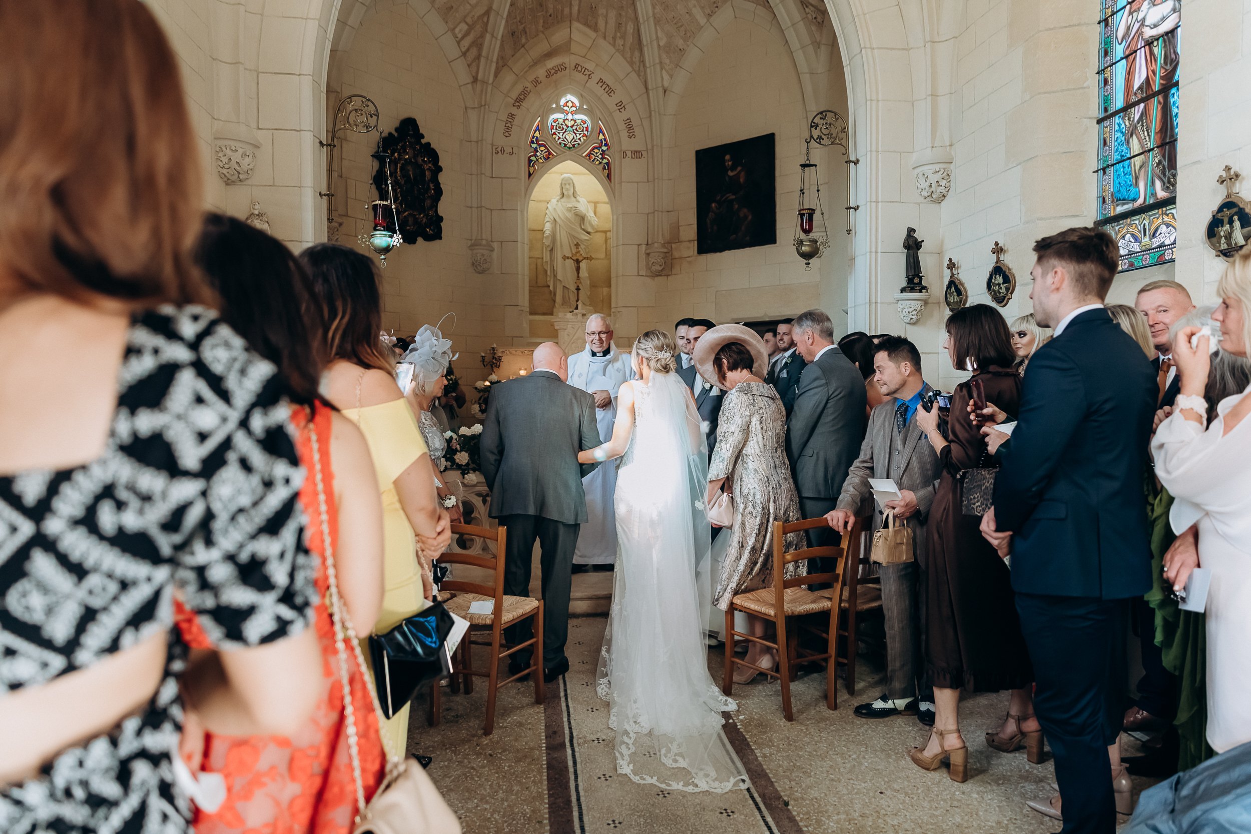 A religious wedding ceremony is taking place inside a small French Catholic chapel with cream limestone walls, Gothic arched stonework, colorful stained glass windows, and a white marble statue of the Virgin Mary in the altar niche. The bride, wearing a fitted ivory lace gown with a long lace-edged veil, stands at the altar alongside a man in a dark grey suit, facing an elderly officiant in white vestments. Approximately 20–25 guests line both sides of the narrow aisle, dressed in colorful formal attire including a yellow dress, red-and-white print dress, and a gold brocade coat with a wide-brim hat. The chapel is furnished with simple rush-seat wooden chairs, and white floral arrangements are visible near the altar. The image is a wide shot taken from the back of the chapel looking toward the altar, with guests filling the foreground on both sides.