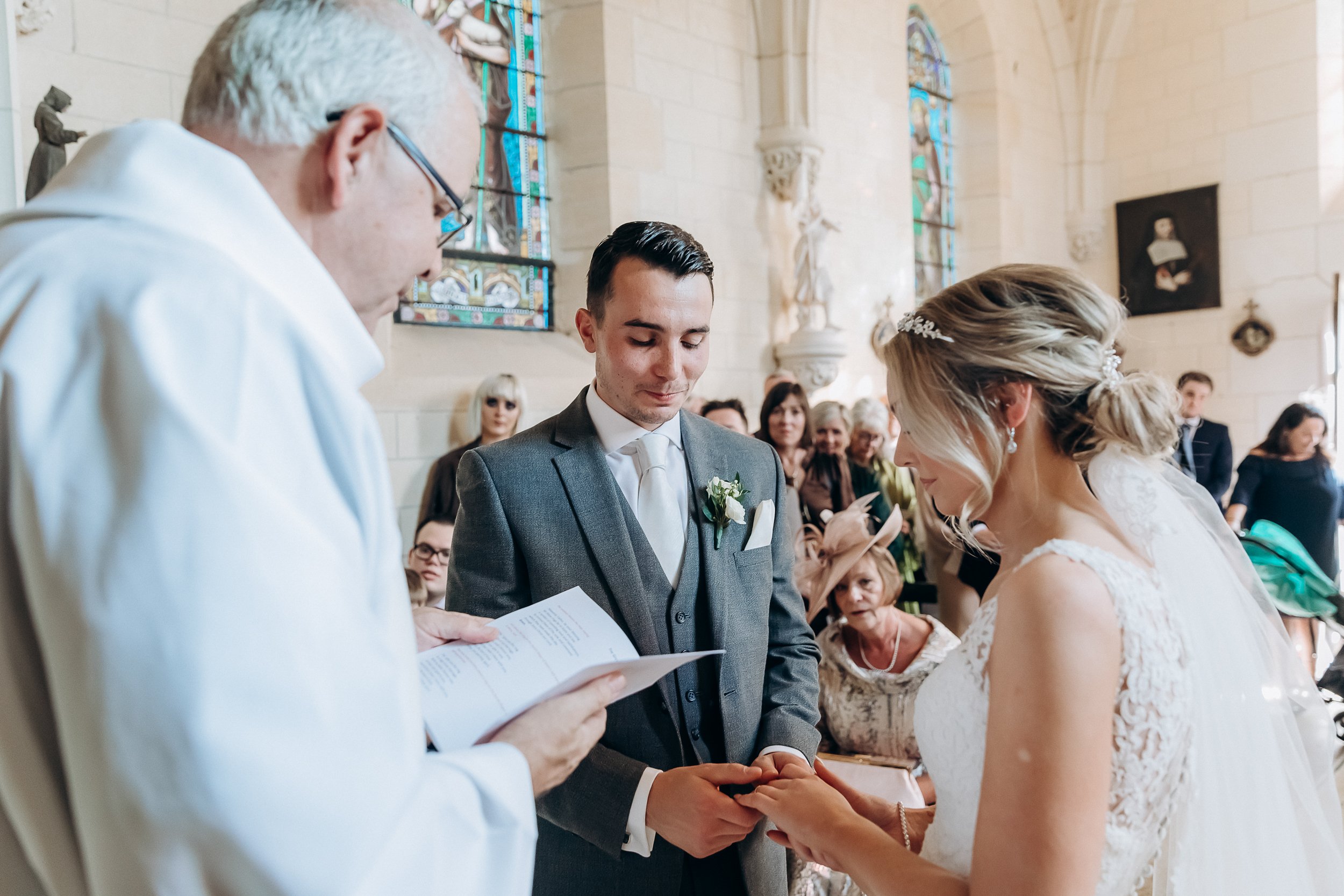 The ring exchange is taking place during an indoor religious ceremony inside a stone-walled chapel or church, featuring colorful stained glass windows, stone columns, and religious statues and paintings on the walls. A priest in white vestments reads from a printed order of service while the groom, wearing a charcoal grey three-piece suit with an ivory tie and a white rose boutonniere, holds the bride's hands as she places a ring on his finger. The bride wears a white lace-detailed gown with a pearl bracelet and a crystal hair comb in her upswept blonde hair. Approximately ten to fifteen seated guests are visible in the pews behind the couple, several wearing formal attire including fascinators in blush and teal tones. The shot is a medium wide portrait taken from a slight side angle, capturing all three principal figures and the congregation in the background.