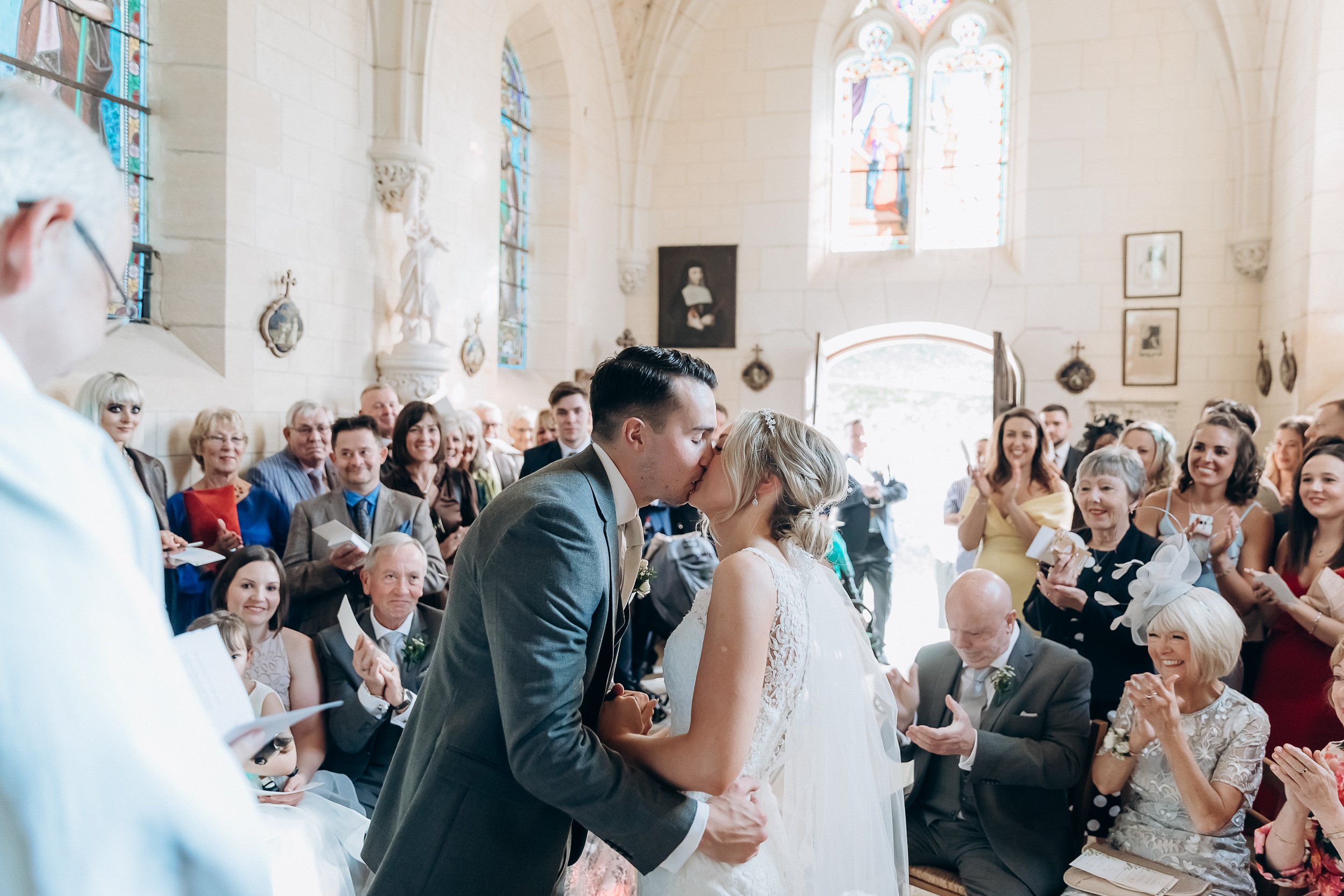 The couple shares their first kiss during an indoor church ceremony in a French chapel with white limestone walls, Gothic arched stained glass windows, and religious artwork and sculptures on the walls. The groom wears a grey suit with a light champagne tie, and the bride wears a white lace-back sleeveless gown with a veil and a small hair accessory, holding what appears to be a small bouquet with greenery. Approximately 30–40 guests fill the wooden pews on both sides of the aisle, many smiling, clapping, and holding confetti; guests are dressed in a mix of formal attire including a yellow dress, red dress, royal blue outfit, and a white fascinator hat. The shot is a wide mid-level perspective taken from the altar end looking back slightly, capturing both the couple in the foreground and the full congregation behind them.