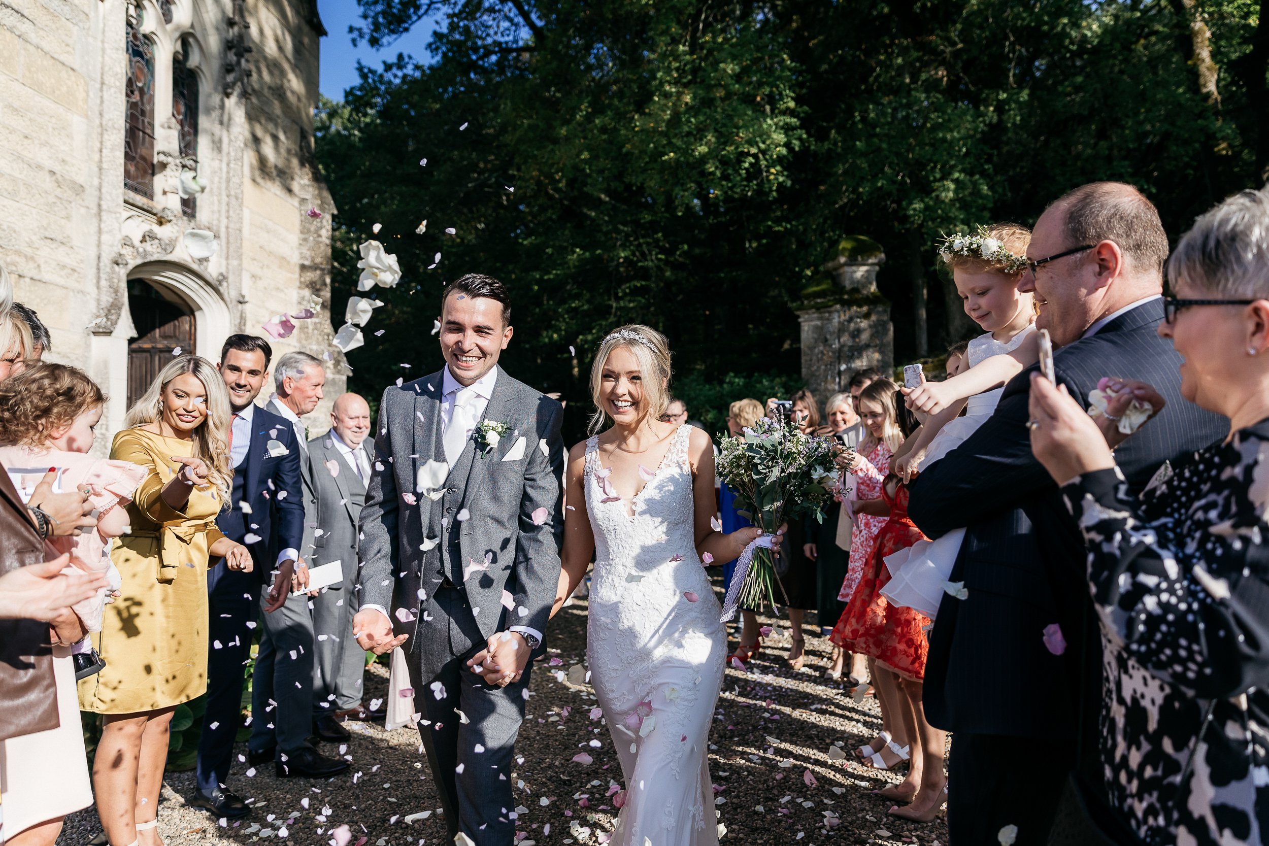 The couple is walking through a confetti exit immediately following their outdoor ceremony, passing through two lines of approximately 20–25 cheering guests outside a stone chapel or church with ornate arched doorways. The groom wears a light grey three-piece suit with a pale blue tie and a white rose boutonniere, while the bride wears a fitted ivory lace gown with a deep V-neckline and carries a loose bouquet of white blooms and greenery with hints of lavender. Pink and white petal confetti fills the air around them as guests throw handfuls from both sides. A young flower girl on the right wears a white and greenery floral crown. The overall styling is classic and polished. Wide-angle documentary-style shot taken in bright midday sunlight.