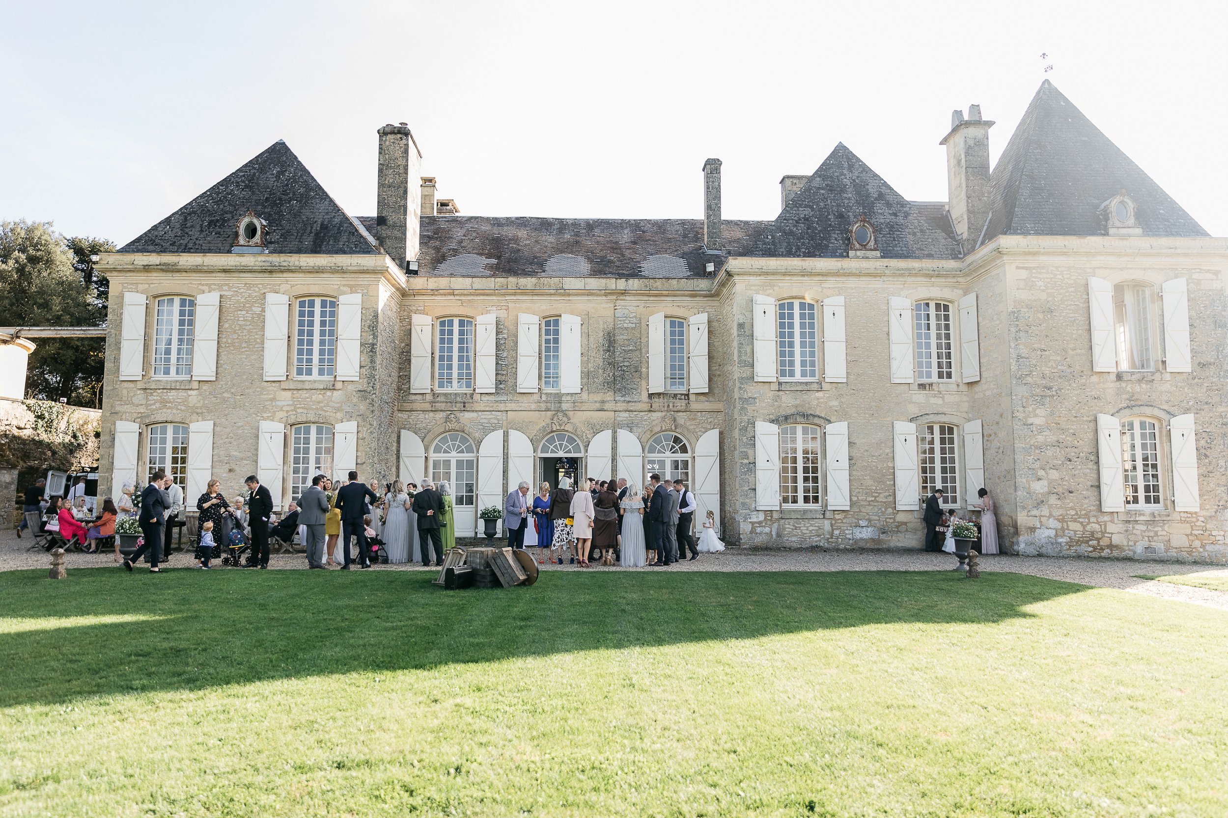 An outdoor cocktail hour is taking place on the gravel terrace and lawn in front of a two-story French chateau with cream limestone facades, dark slate pointed rooftops, and white shuttered windows. Approximately 40–50 guests are mingling in small groups across the front of the building, dressed in a mix of formal attire including dark suits, colourful occasion dresses in mustard, teal, red, and floral prints, and light grey bridesmaid gowns. The bride in a white gown is visible near the central entrance alongside the wedding party. A rustic wooden crate prop sits on the gravel in the foreground. The shot is a wide exterior view taken from across the lawn, capturing both the full chateau facade and the crowd gathered before it. Potential venue feature image.