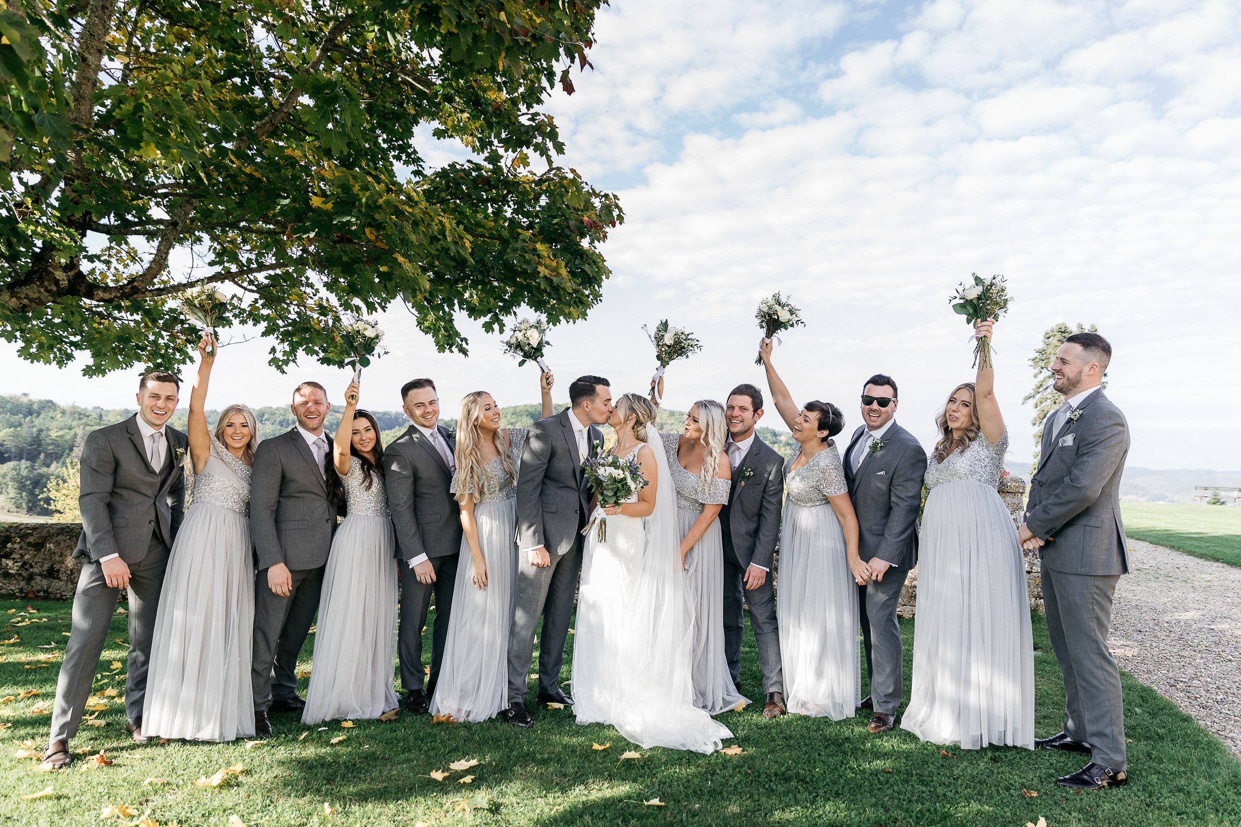 A bridal party group portrait taken outdoors on a lawn with a panoramic countryside view in the background. The bride, in a white sleeveless A-line gown with a veil, kisses the groom at the center of the group; the groom and five groomsmen are dressed in charcoal grey suits with blush/mauve ties and boutonnieres. Five bridesmaids wear floor-length silver-grey dresses with sequined or beaded bodices, and several raise their bouquets of white flowers and greenery into the air in celebration. The overall styling palette is silver-grey, charcoal, and white with greenery accents, giving a modern classic feel. The wide shot captures all 12 members of the bridal party laughing and cheering, set against a gravel pathway and low stone wall to the right.