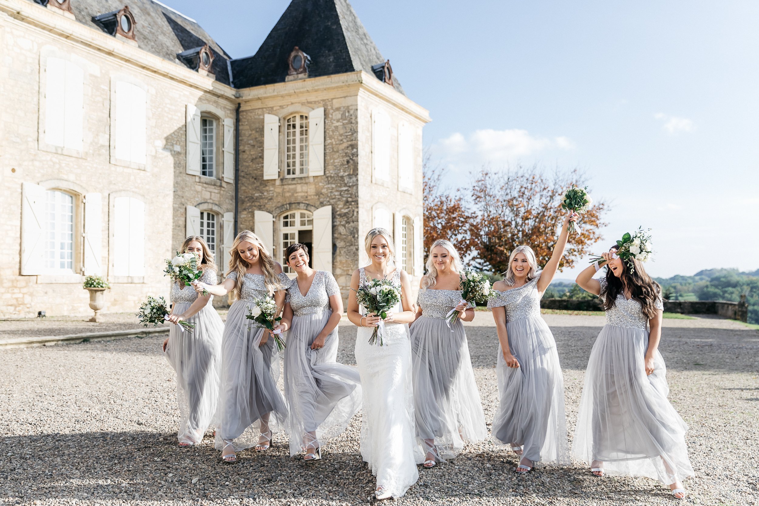 A bridal party portrait taken outdoors on the gravel forecourt of a French chateau, with the limestone facade and white shuttered windows prominent in the background. The bride stands centrally in a fitted white lace gown with a beaded headpiece, surrounded by six bridesmaids wearing floor-length silver-grey tulle dresses with sequined bodices in varying neckline styles. All seven women are walking toward the camera in a relaxed, candid manner — laughing and smiling, with one bridesmaid on the far right raising her bouquet in the air. Each bridesmaid carries a bouquet of white roses and ivory blooms with lush greenery, matching the bride's own bouquet. The styling palette is cohesive in silver, white, and green, with a classic-meets-modern aesthetic. Wide shot composition capturing the full group and the chateau architecture. Potential venue feature image.