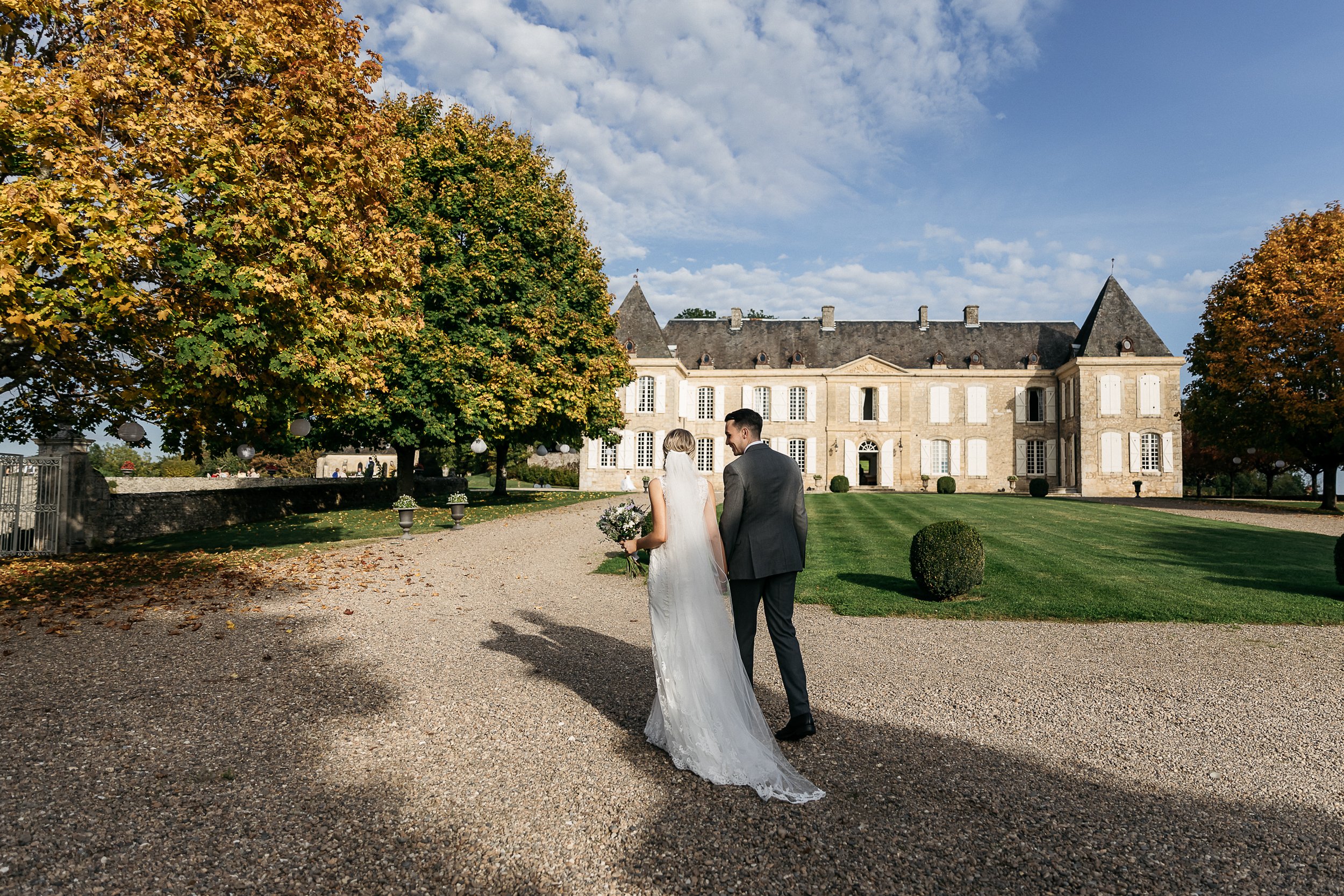 A couple portrait taken outdoors on the gravel forecourt of a French château, shot from behind in a wide composition. The bride wears a white lace gown with a long cathedral-length veil and carries a small bouquet with white blooms and greenery; the groom wears a charcoal grey suit. The two stand close together, with the groom turning slightly toward the bride. The château in the background is a classic French manor house in honey-toned stone with a grey slate roof, white shutters, and symmetrical façade, framed by mature trees showing autumn gold and orange foliage. A small group of guests is visible in the mid-distance near the entrance gates. Potential venue feature image.