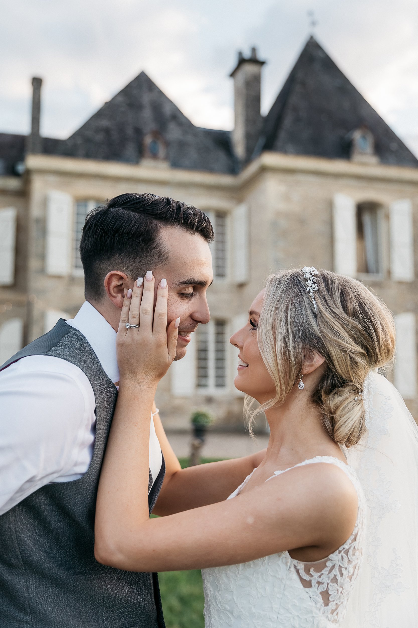 A couple portrait taken outdoors in front of a French chateau with stone facade and steeply pitched slate rooftops. The bride and groom face each other closely, with the bride placing her hand gently against the groom's cheek, her diamond engagement ring and wedding band clearly visible. The groom wears a charcoal grey waistcoat over a white dress shirt with a dark tie, while the bride wears a lace wedding gown with illusion back detailing and thin straps. Her blonde hair is styled in a low updo accessorized with a crystal floral hair comb, and she wears teardrop crystal earrings. The composition is a close-up portrait with the chateau softly blurred in the background.