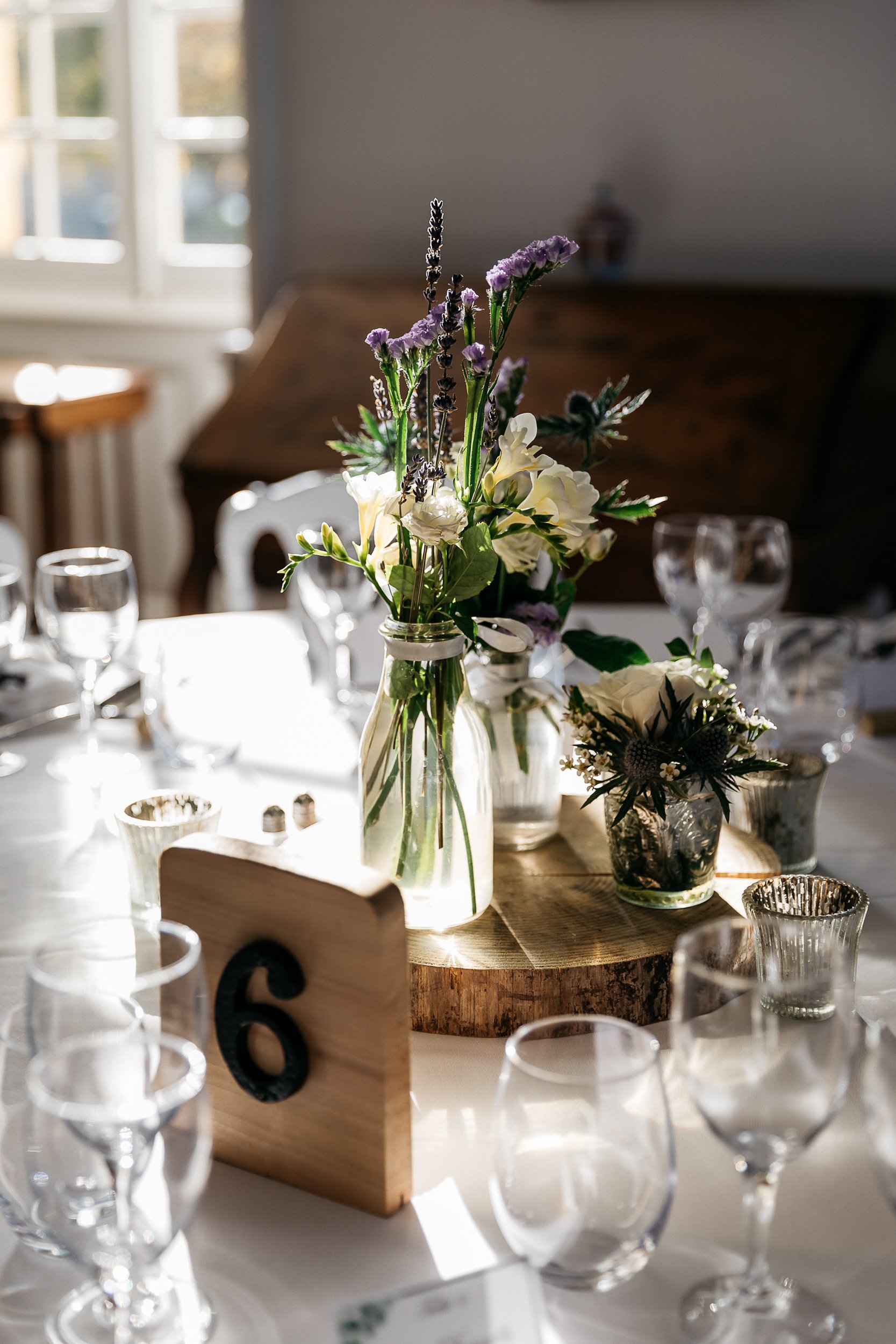 Close-up detail shot of a wedding reception table centerpiece with a rustic styling theme. The centerpiece sits on a raw wood slice and features small glass bud vases holding an informal wildflower arrangement of white ranunculus, white freesia, purple lavender, purple statice, blue eryngium thistles, and mixed greenery, alongside small mercury glass votive candle holders. A wooden block table number marker displaying the number 6 stands in front of the arrangement. The white linen-covered table is set with multiple wine glasses and water glasses, with additional tables and white chairs visible in the softly blurred background of what appears to be an indoor reception room with natural light coming through white-framed windows.