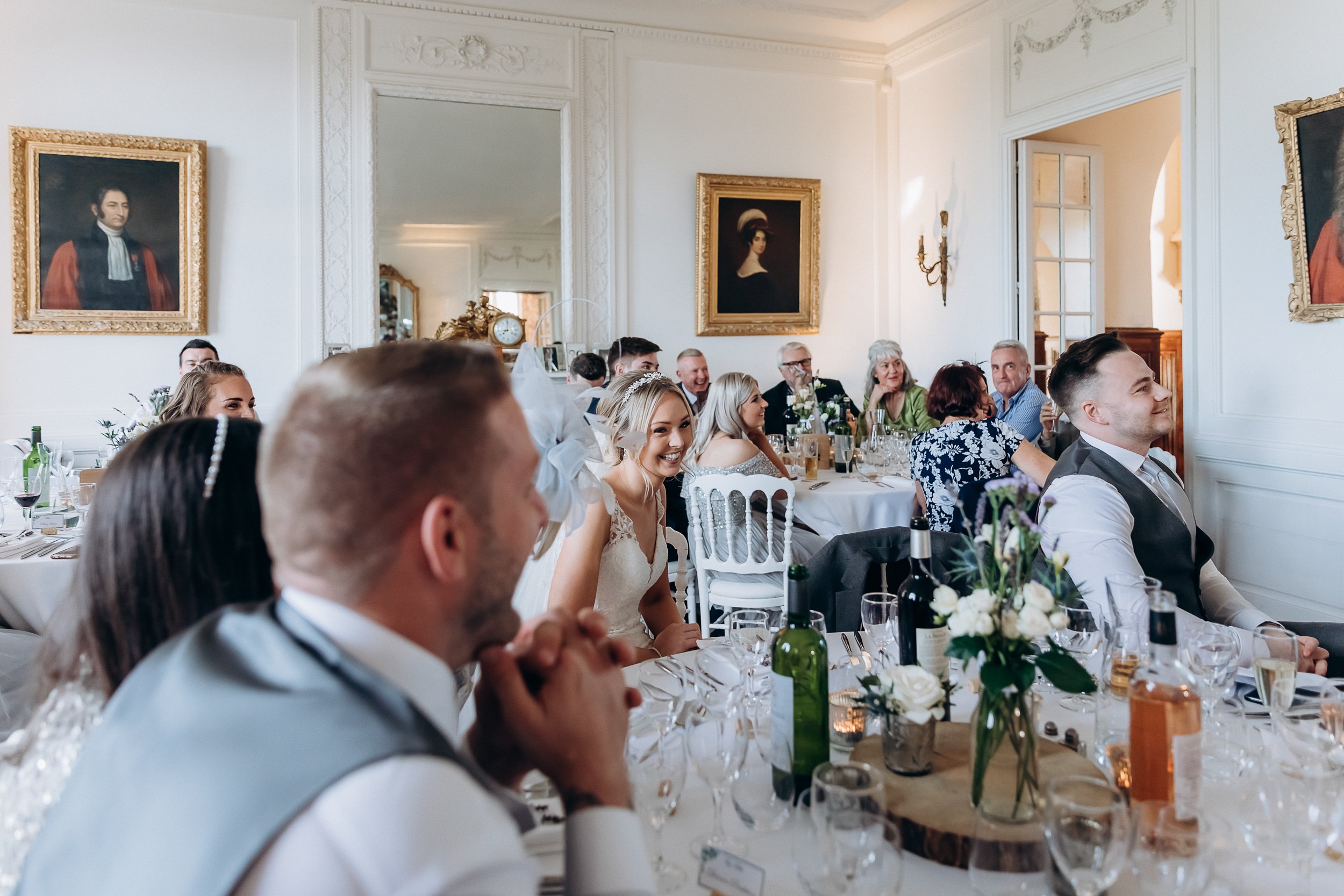 Wedding reception speeches captured inside a classic chateau or manor house dining room, with approximately 20 guests seated at white linen-covered tables listening and laughing. The bride, wearing a lace fitted gown with a floral hairpiece, is seated mid-table and smiling broadly, while the groom in the foreground wears a pale blue waistcoat. The room features white panelled walls with ornate gold-framed oil portrait paintings, a large gilt mirror, and wall sconce lighting, giving the setting a classic French château aesthetic. Table centrepieces consist of white roses and purple-toned flowers in glass vases on wooden slice bases, with wine bottles, glassware, and small candles completing the table styling. The image is a wide environmental shot taken from a low angle at the head table end, capturing the full depth of the room.
