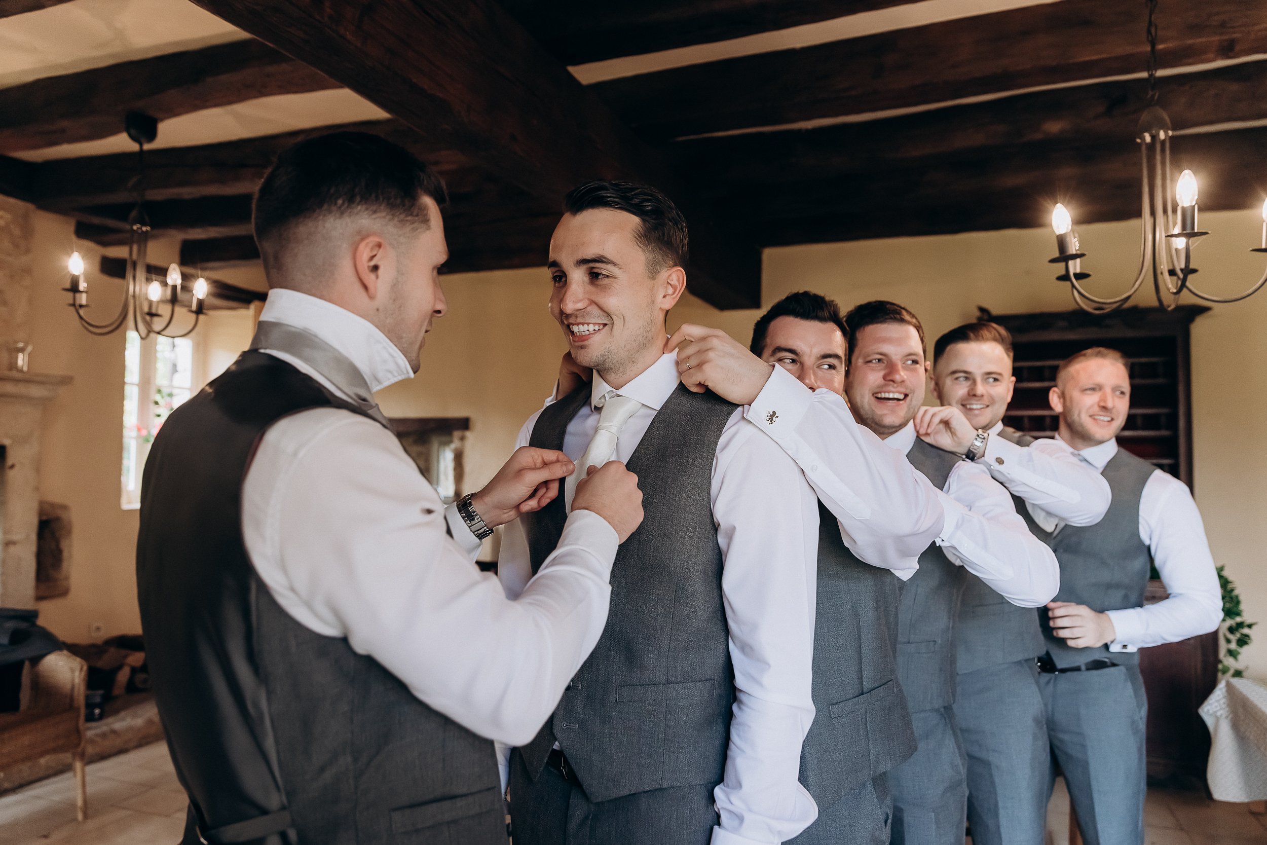 The groom and his groomsmen are getting ready indoors in what appears to be a historic venue room with dark exposed wooden beam ceilings and wrought-iron candle-style chandeliers. Five men are visible, all wearing grey waistcoats over white dress shirts with grey trousers; the groom in the center is having his ivory tie adjusted by one groomsman while others playfully help straighten his collar, all laughing. The groom's cufflinks are visible — gold-toned with a crest detail. The setting has warm ambient lighting from the chandeliers and features a stone fireplace in the background, giving the room a classic, rustic interior feel. This is a candid mid-shot capturing a lighthearted getting-ready moment.