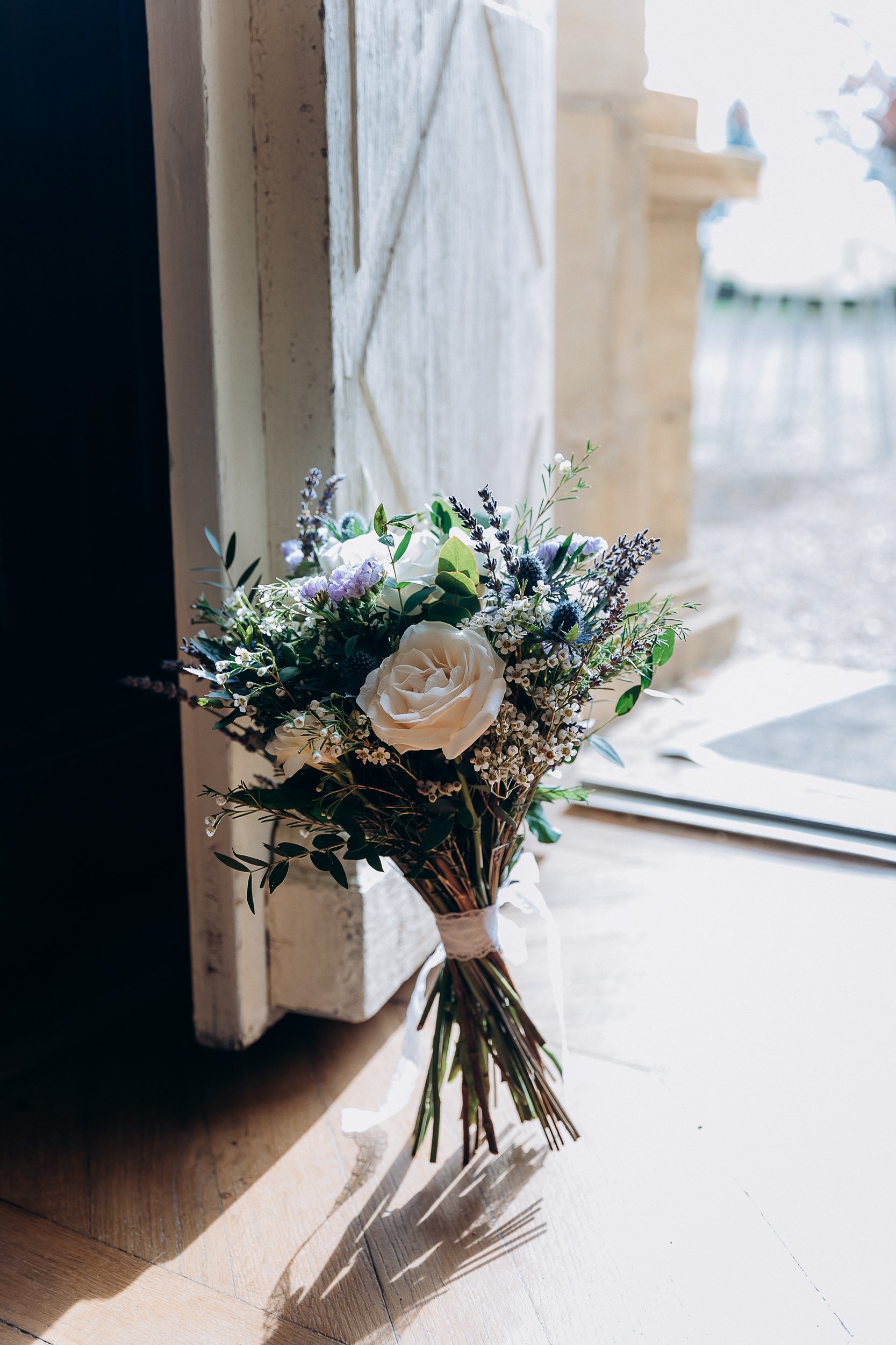 Close-up detail shot of a bridal bouquet resting upright against a white-painted wooden door frame on a light wood floor, with natural window light casting soft shadows. The bouquet features cream garden roses, blue thistle, lavender, white waxflower, and mixed greenery including eucalyptus and olive-style foliage, tied with a white ribbon. The loose, wildflower-style arrangement has a rustic, garden-gathered aesthetic. A stone balustrade is faintly visible through the window in the background, suggesting a French chateau or manor setting.