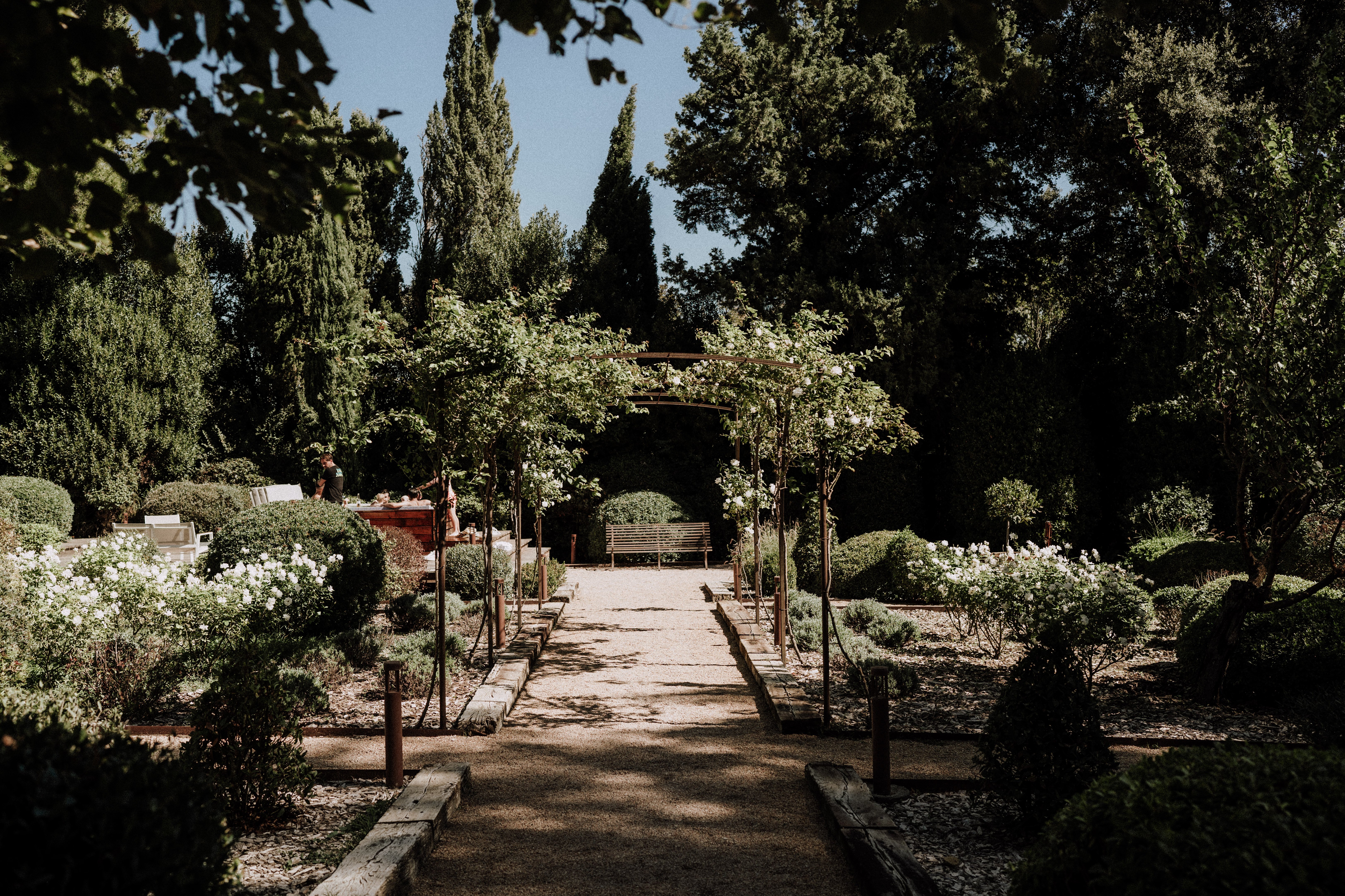 A wide shot of a formal French garden being set up for a wedding, captured in natural daylight. The scene features a central gravel pathway leading through a wooden pergola structure draped with climbing foliage and white roses, flanked by neatly clipped box hedges, topiary, and white rose bushes on either side. A wooden garden bench sits at the far end of the path near a hedge archway, and low wooden post lighting lines the walkway. To the left, one or two people appear to be arranging items at a dark red/wooden bar or service table, suggesting pre-event setup is underway. Potential venue feature image.