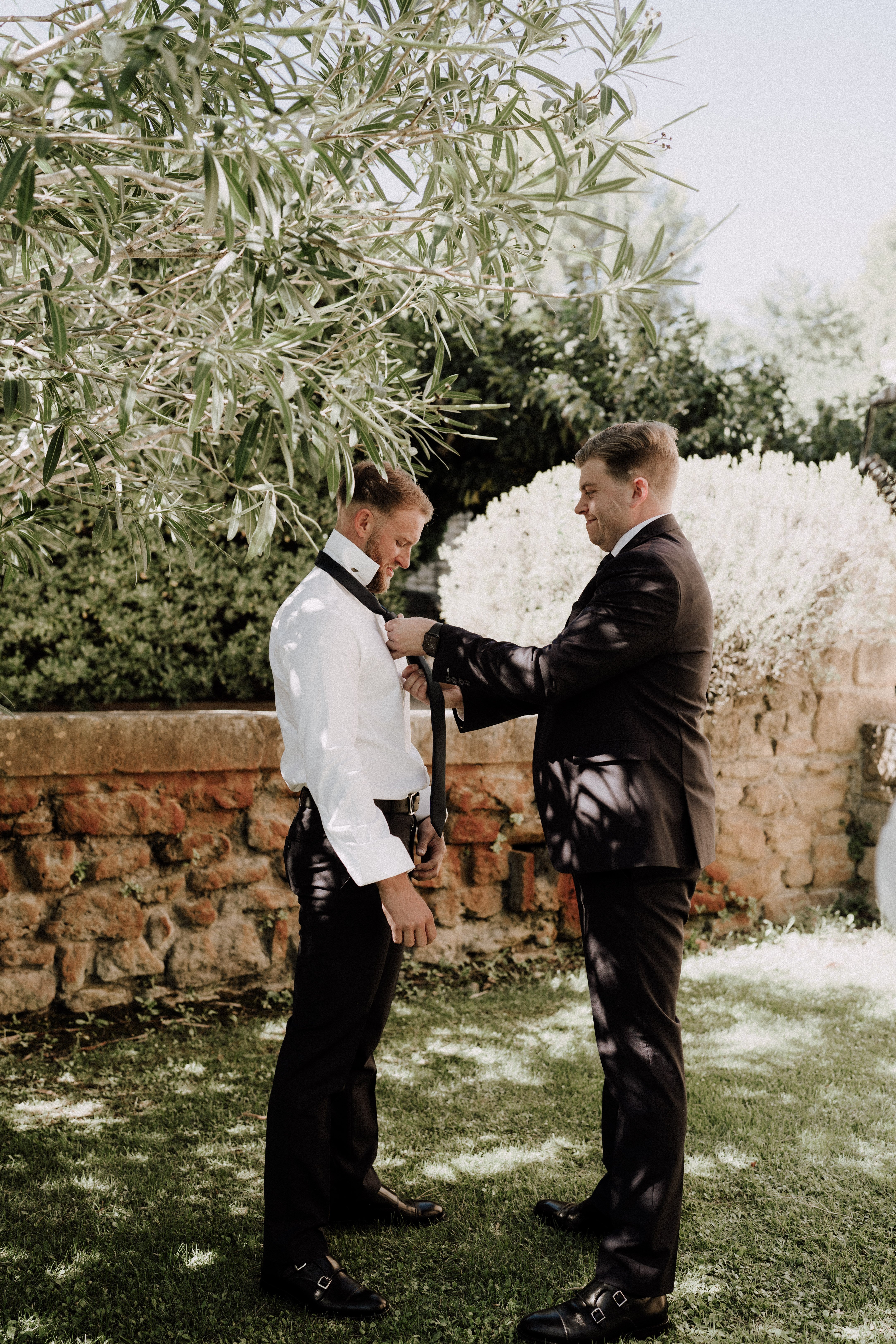 A getting-ready moment captured outdoors in a garden setting, where one man in a white dress shirt and black trousers helps another man in a dark navy suit adjust a black tie. Both men wear black dress shoes — the groom in monk-strap style. The setting features a rustic exposed brick and stone garden wall in the background, with an arched opening visible, suggesting a chateau or domaine property. The shoot is a full-length portrait taken in natural daylight with dappled shade from an olive tree overhead.