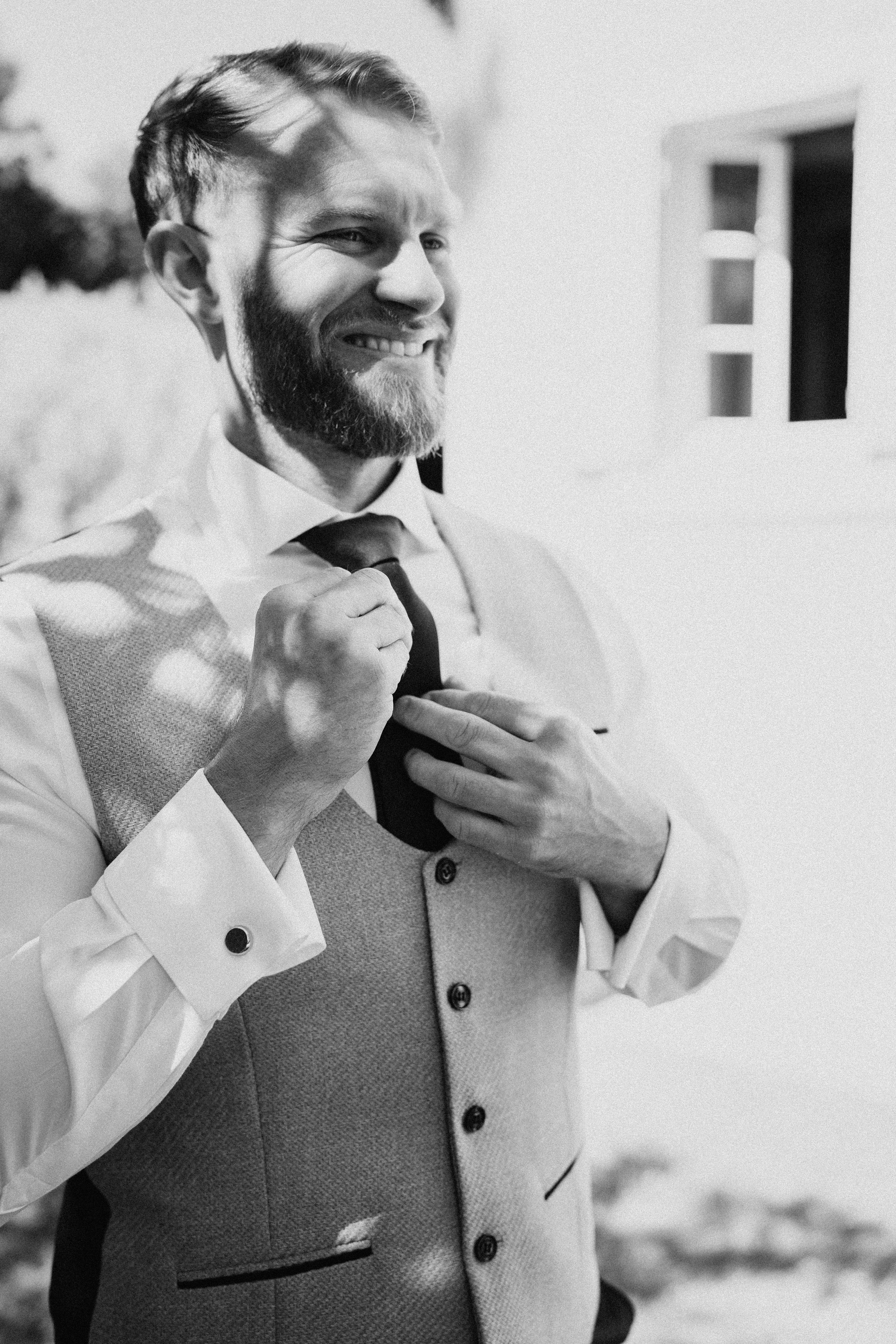 This black-and-white portrait captures a groom during the getting-ready phase, adjusting his tie while smiling broadly. He is dressed in a textured waistcoat, a white dress shirt with French cuffs and visible cufflinks, and a dark tie. The image is a close-up portrait shot with shallow depth of field, taken outdoors against a light-rendered building facade. High contrast tones highlight the dappled light falling across his waistcoat and shirt.