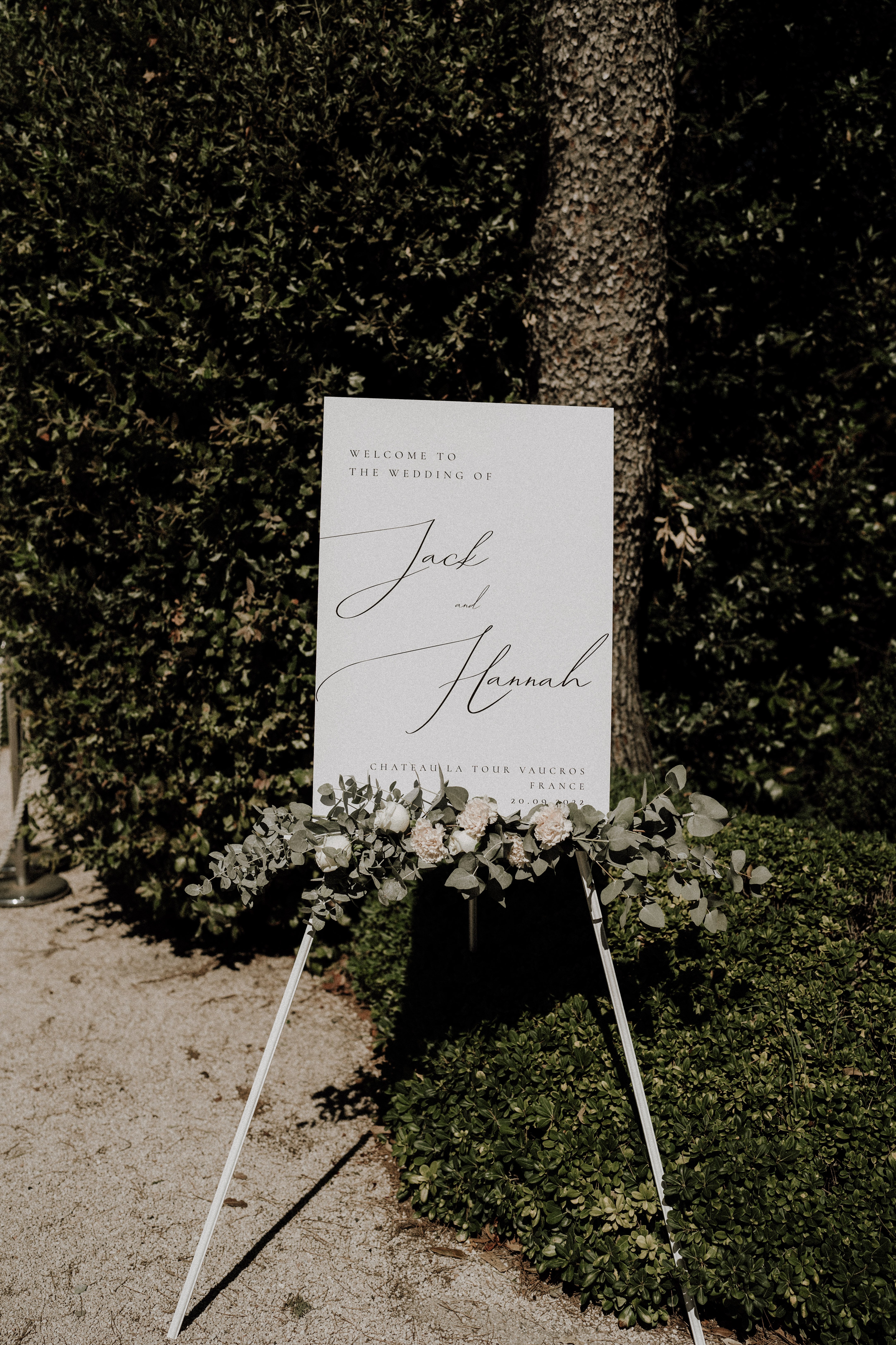 A close-up detail shot of a white rectangular welcome sign displayed on a white metal easel outdoors at Chateau La Tour Vaucros, France. The sign reads 'Welcome to the Wedding of Jack and Hannah' in a combination of small-caps serif and large script typography, with the venue name and date (20.09.2022) printed at the bottom. The base of the easel is decorated with a cluster of eucalyptus sprigs, white roses, and blush carnations or garden roses arranged in a loose, organic style. The sign is positioned against a trimmed hedge and a tree trunk in a gravel garden setting, with bright dappled sunlight creating strong shadows. The overall decor palette is white and green with a modern minimalist aesthetic.