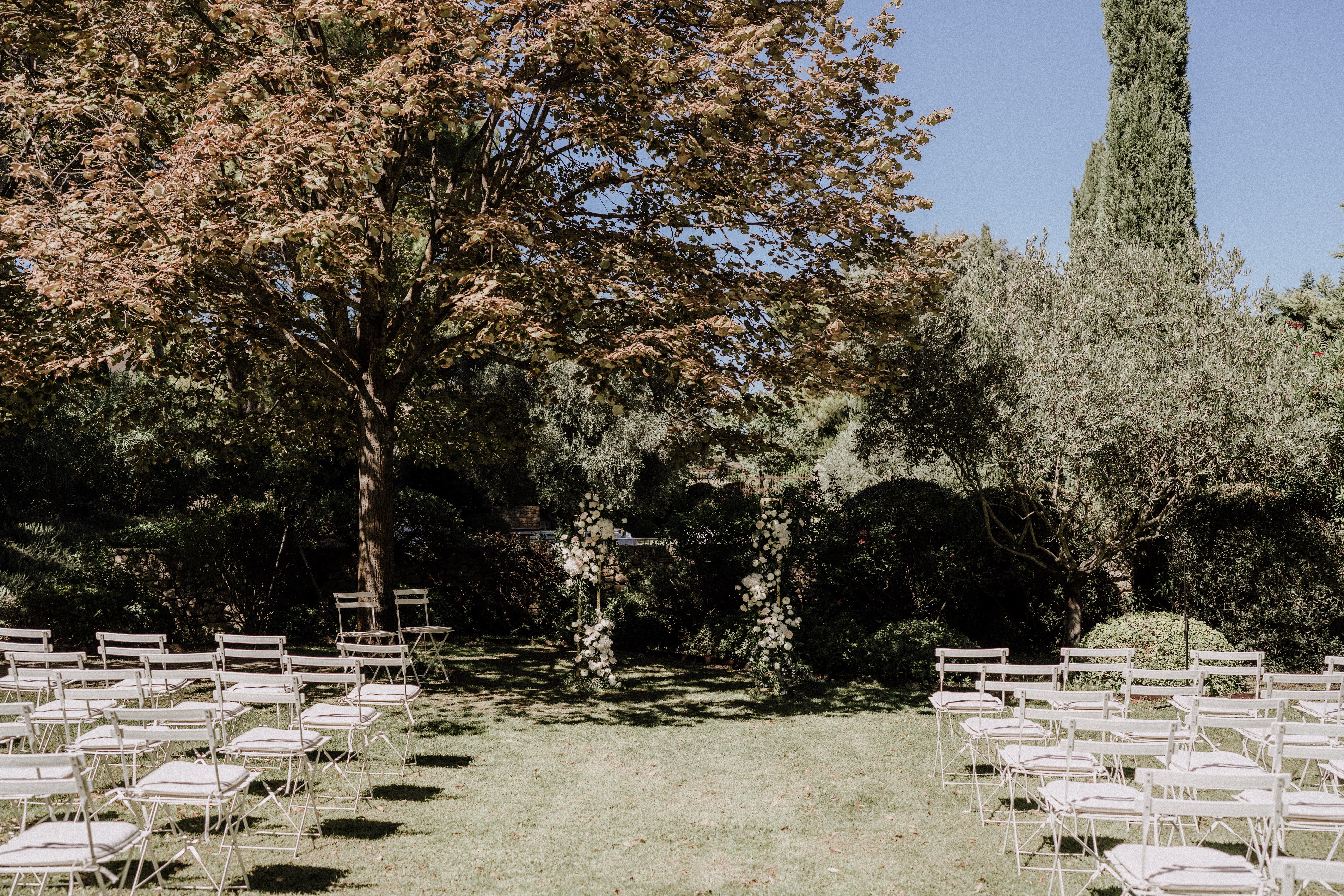 An outdoor wedding ceremony setup photographed before guests arrive, set on a lawn within a garden. Rows of light grey metal folding chairs with white cushioned seat pads are arranged on either side of a central aisle, facing a circular floral arch decorated with white blooms and trailing greenery, likely eucalyptus. The decor palette is white and green, giving the ceremony space a clean, minimalist feel. Wide shot capturing the full ceremony layout with no people present.