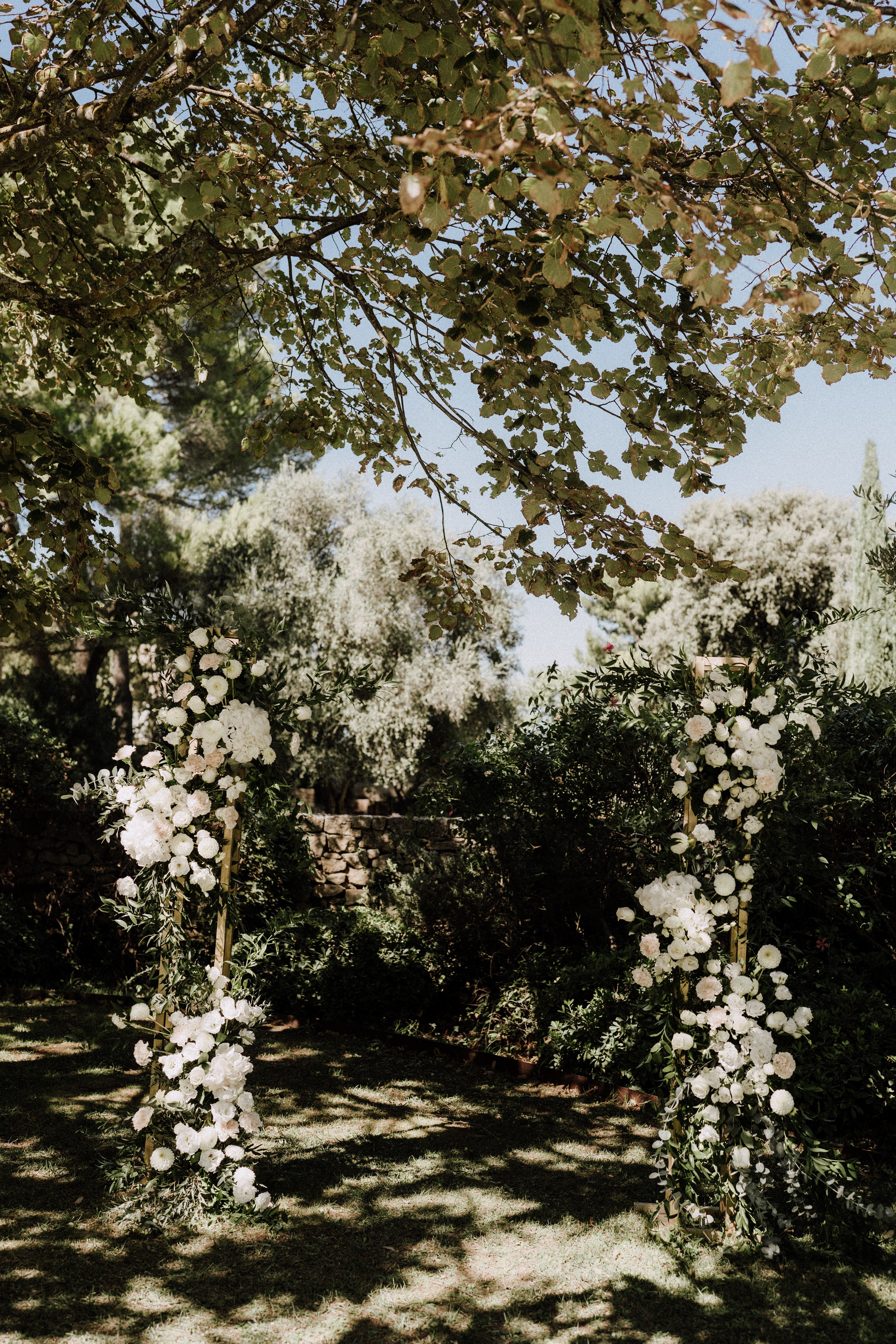 An outdoor ceremony setup featuring a square gold metal arch decorated with cascading clusters of white peonies, hydrangeas, ranunculus, and trailing green foliage on both uprights. No people are present — this is a detail shot of the ceremony backdrop set in a garden, with a low stone wall visible in the background and dense greenery surrounding the space. The floral palette is entirely white and ivory with deep green leaves, creating a classic, garden-style aesthetic. The image is a wide portrait-orientation shot taken in natural daylight with dappled shade from an overhanging tree.