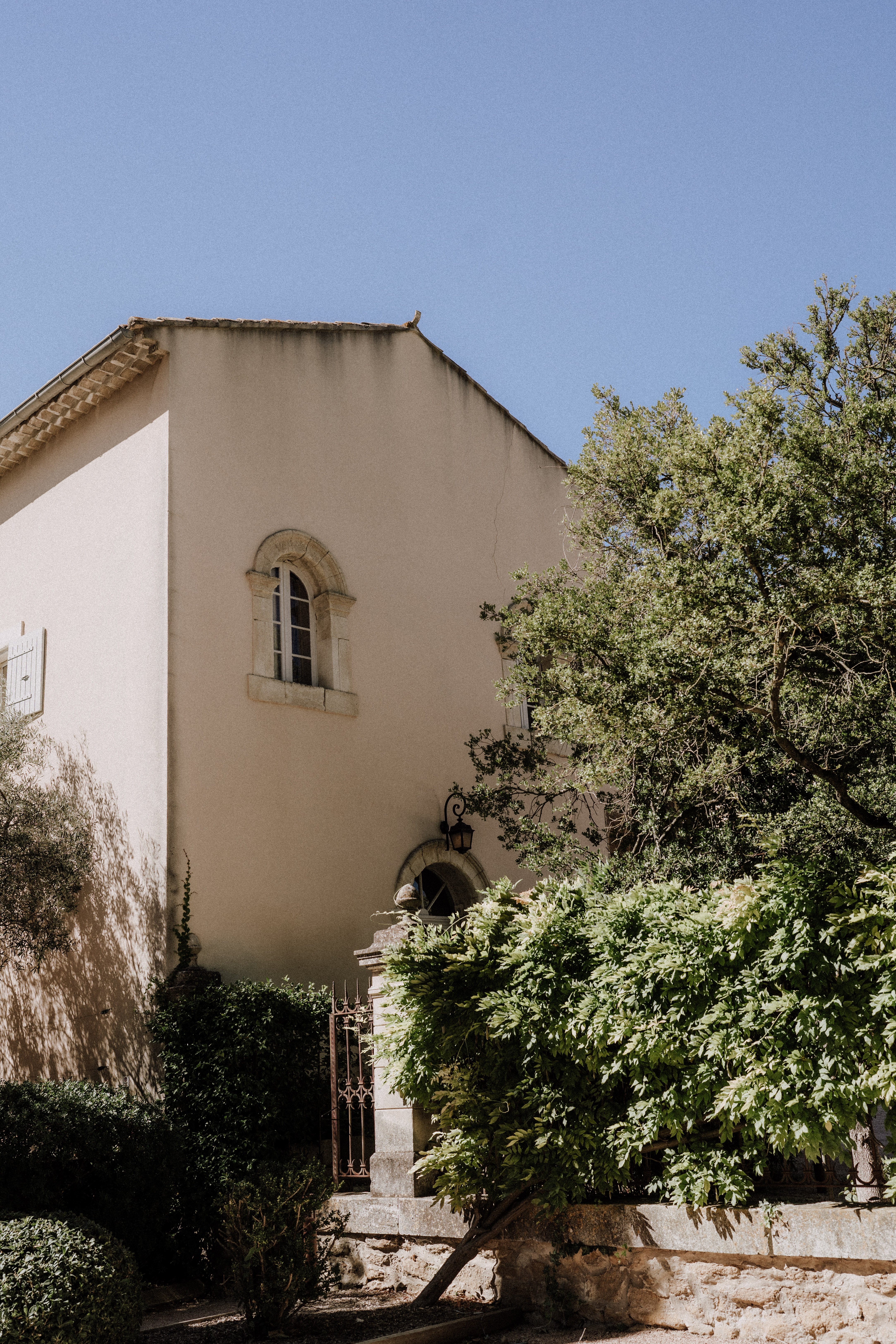 An outdoor wide shot of a French country manor or mas, featuring warm cream-toned rendered walls, a terracotta-tiled roof, and an arched stone-framed window on the upper level. A wrought-iron gate with ornate detailing leads to an entrance porch fitted with a black iron wall lantern and an arched doorway. No people are visible in the frame. Potential venue feature image.