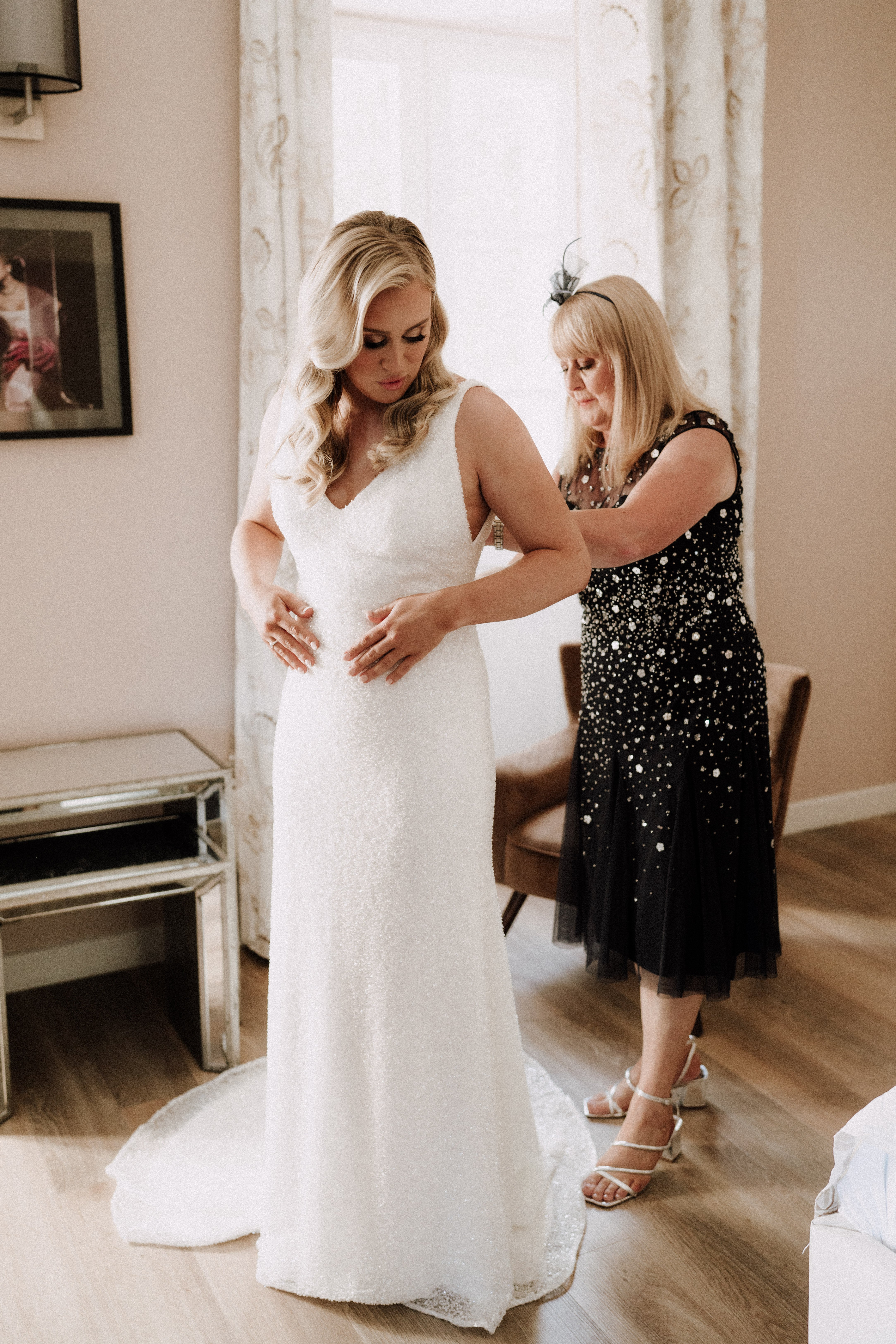 A getting-ready scene captured indoors in what appears to be a hotel room or bridal suite, with warm neutral walls, a mirrored side table, and sheer patterned curtains providing soft backlight. The bride, a blonde woman with loose waves, wears a fitted ivory sequined gown with a V-neckline and a short train, and white strappy block-heeled sandals, while she looks downward as a second woman fastens the back of the dress. The second woman, likely the mother of the bride, wears a knee-length black embellished dress with scattered pearl and sequin detailing and a small dark fascinator headpiece. The composition is a full-length portrait shot of the two women, with a framed photograph visible on the wall to the left.