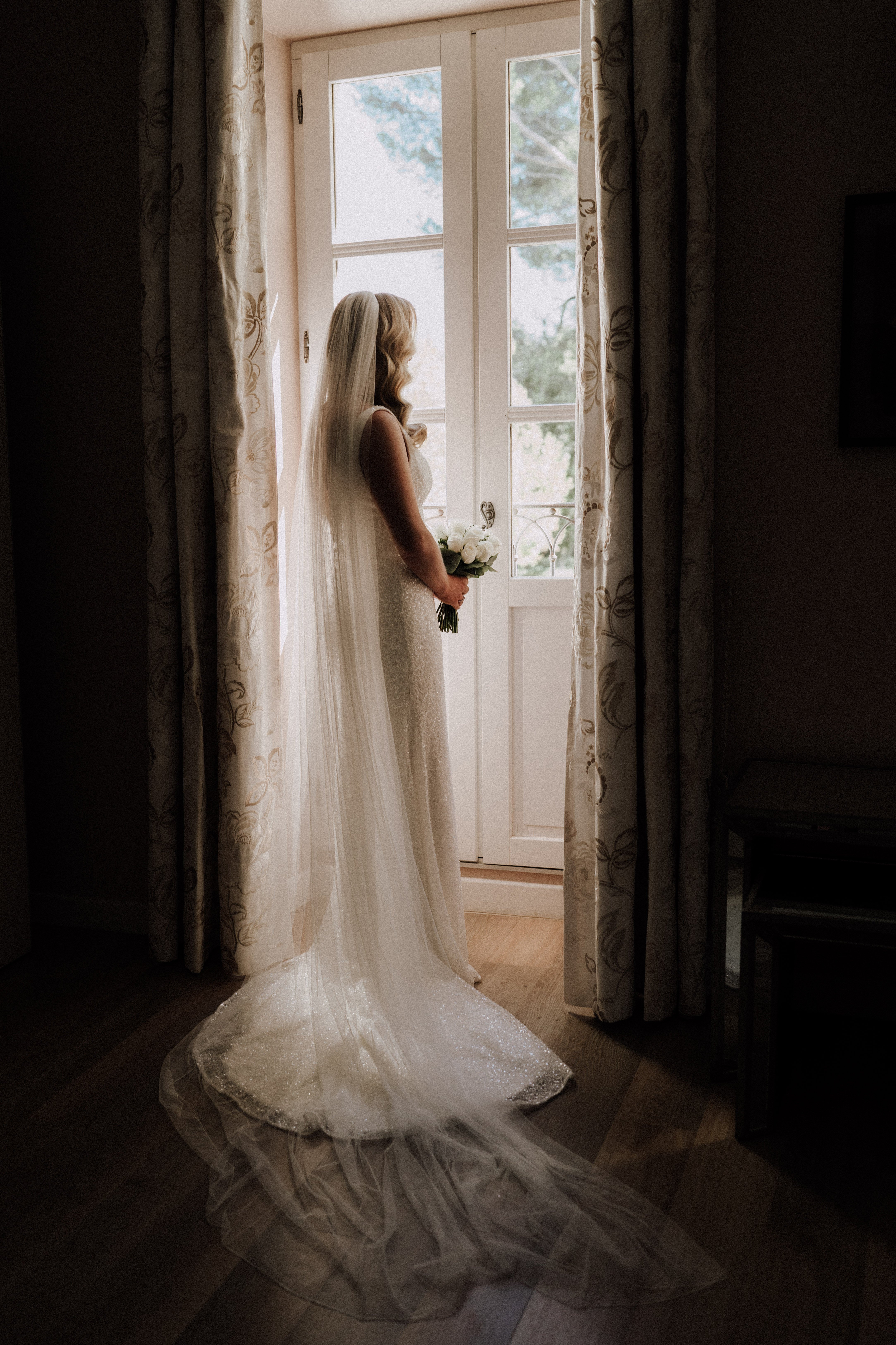 A bridal portrait taken indoors, showing the bride from behind as she stands at a set of open French doors, looking outside. She wears a fitted ivory gown with subtle shimmer and a long cathedral-length veil that pools on the hardwood floor, paired with a compact bouquet of white roses. The room features floral-patterned curtains in cream and taupe framing the doorway, creating a strong backlit contrast between the bright exterior and the darker interior. The composition is a full-length portrait emphasizing the back detail of the gown and the dramatic sweep of the veil.