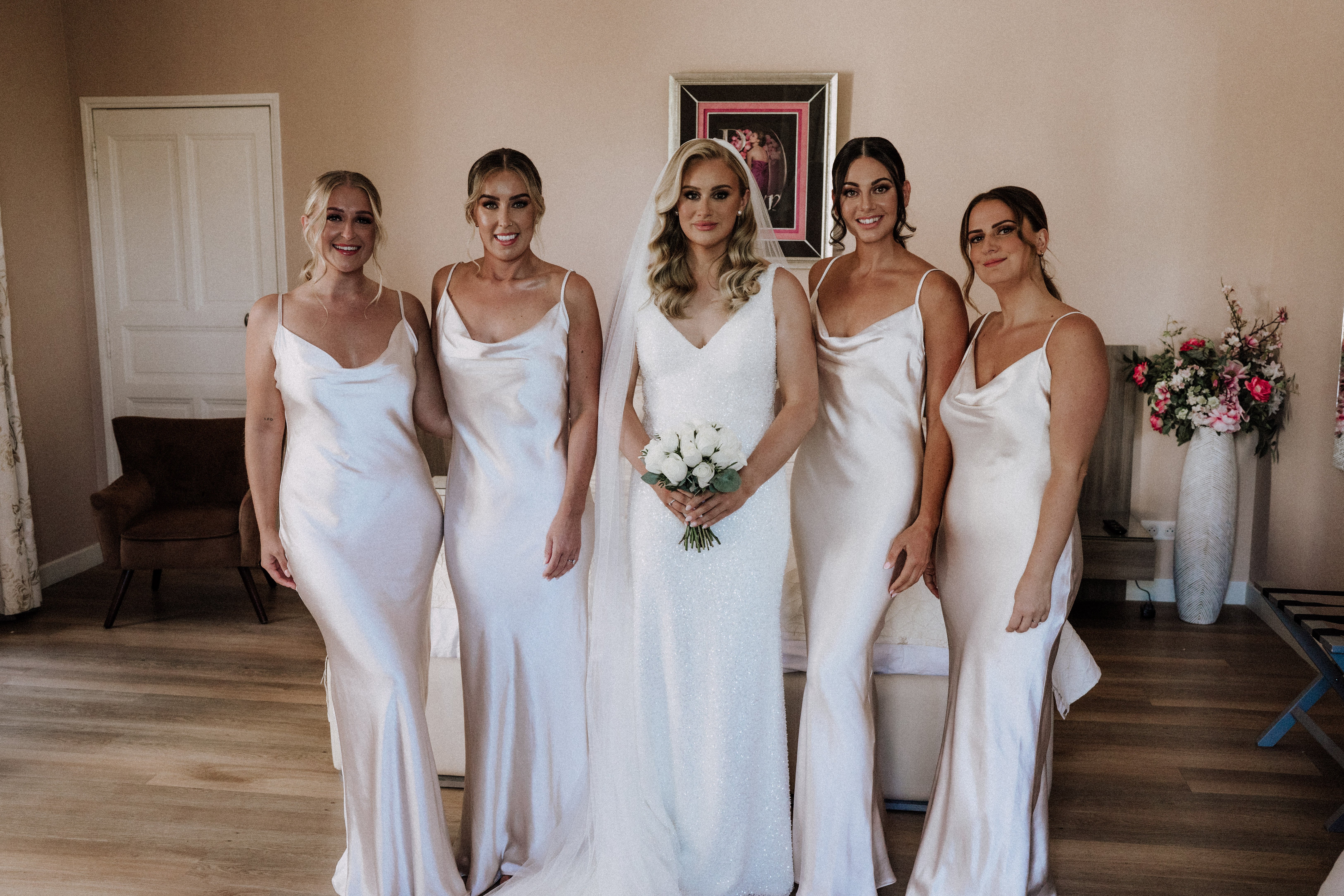 A getting-ready or pre-ceremony portrait of a bride and four bridesmaids posed together indoors in a room with warm beige walls and light wood flooring. The bride wears a form-fitting, fully sequined white gown with a V-neckline and a cathedral-length veil, and holds a compact bouquet of white roses with eucalyptus. The four bridesmaids wear matching champagne satin slip dresses with cowl necklines and spaghetti straps in a sleek, modern style. All five women have their hair styled in soft waves or low updos, with full glam makeup. A tall white textured vase with pink and blush floral arrangements is visible in the background to the right, alongside a framed pink-matted artwork on the wall. This is a mid-length group portrait shot.