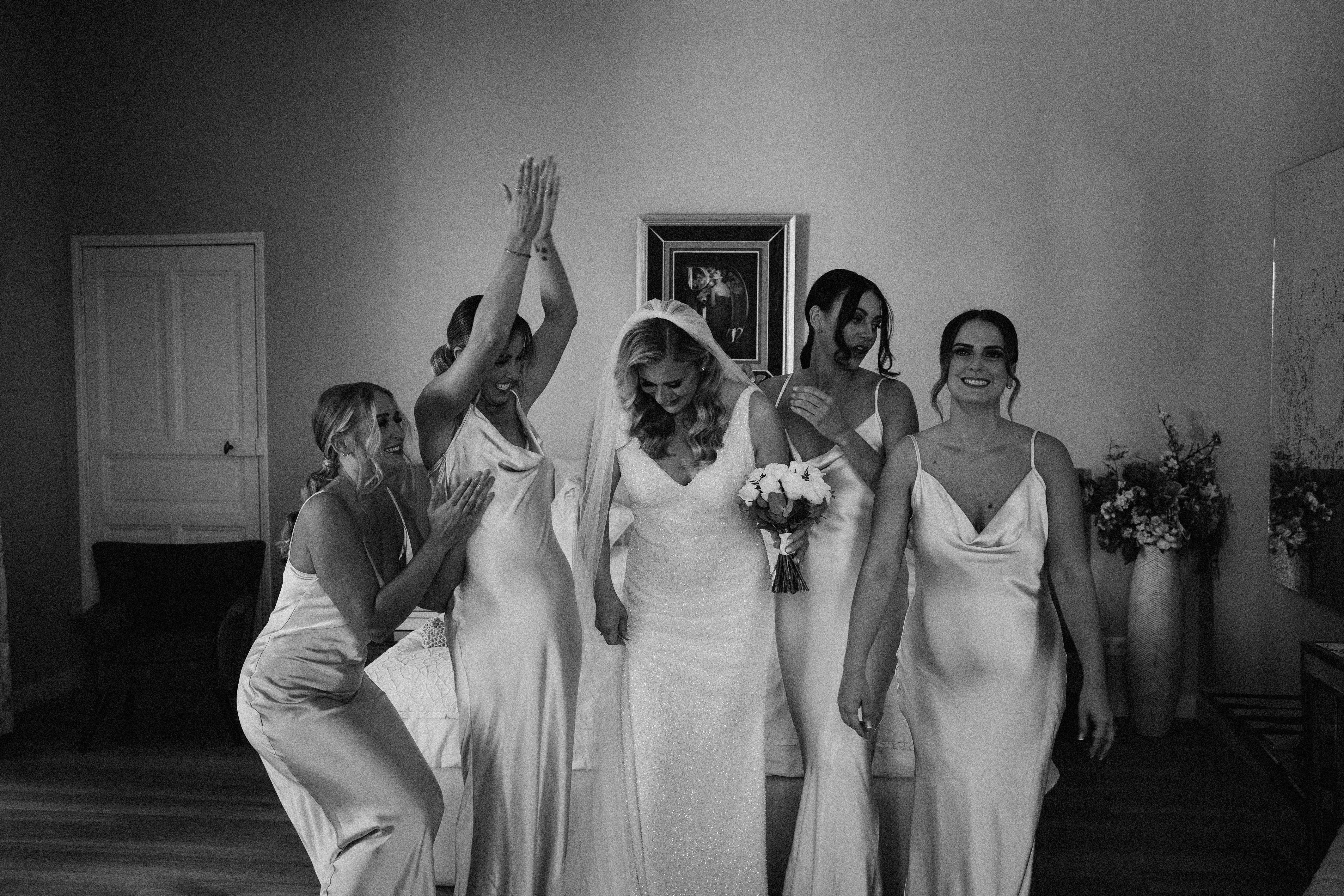 A black-and-white getting-ready moment captured indoors in what appears to be a bedroom or bridal suite, showing the bride surrounded by four bridesmaids in a candid, celebratory pose. The bride wears a fitted, textured white gown with a V-neckline and a cathedral-length veil, and holds a compact bouquet of light-toned flowers. The four bridesmaids wear matching satin slip dresses with cowl or V-necklines, with one bridesmaid raising both arms overhead in excitement while the others gather closely around the bride, laughing and smiling. The image is shot in high contrast black and white, with bright highlights on the dresses against a softly lit plain wall with a framed artwork hanging behind the group; a floral arrangement is partially visible to the right. The composition is a wide portrait shot taken at a slight low angle, emphasizing the energy and movement of the group.