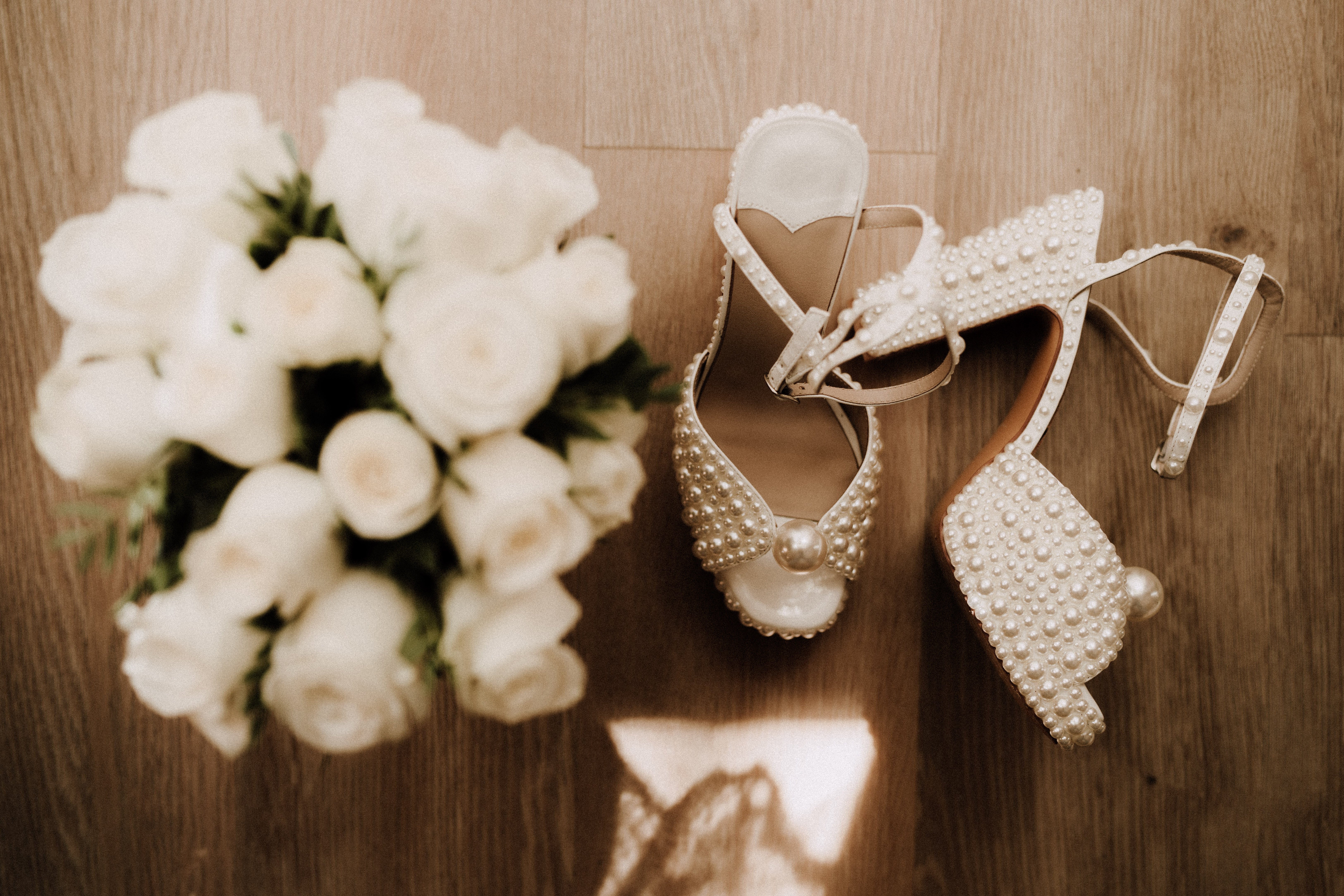 A flat lay detail shot of bridal accessories arranged on a light wood floor, shot from above. The focal point is a pair of pearl-encrusted platform heeled sandals with ankle straps — the shoes are covered densely in ivory and cream pearls of varying sizes, with a large single pearl accent at the toe. To the left, slightly out of focus, is a round bridal bouquet composed of white roses with minimal greenery. A piece of white lace fabric is partially visible at the bottom center of the frame. The overall styling is classic and polished, with a white and ivory color palette throughout.