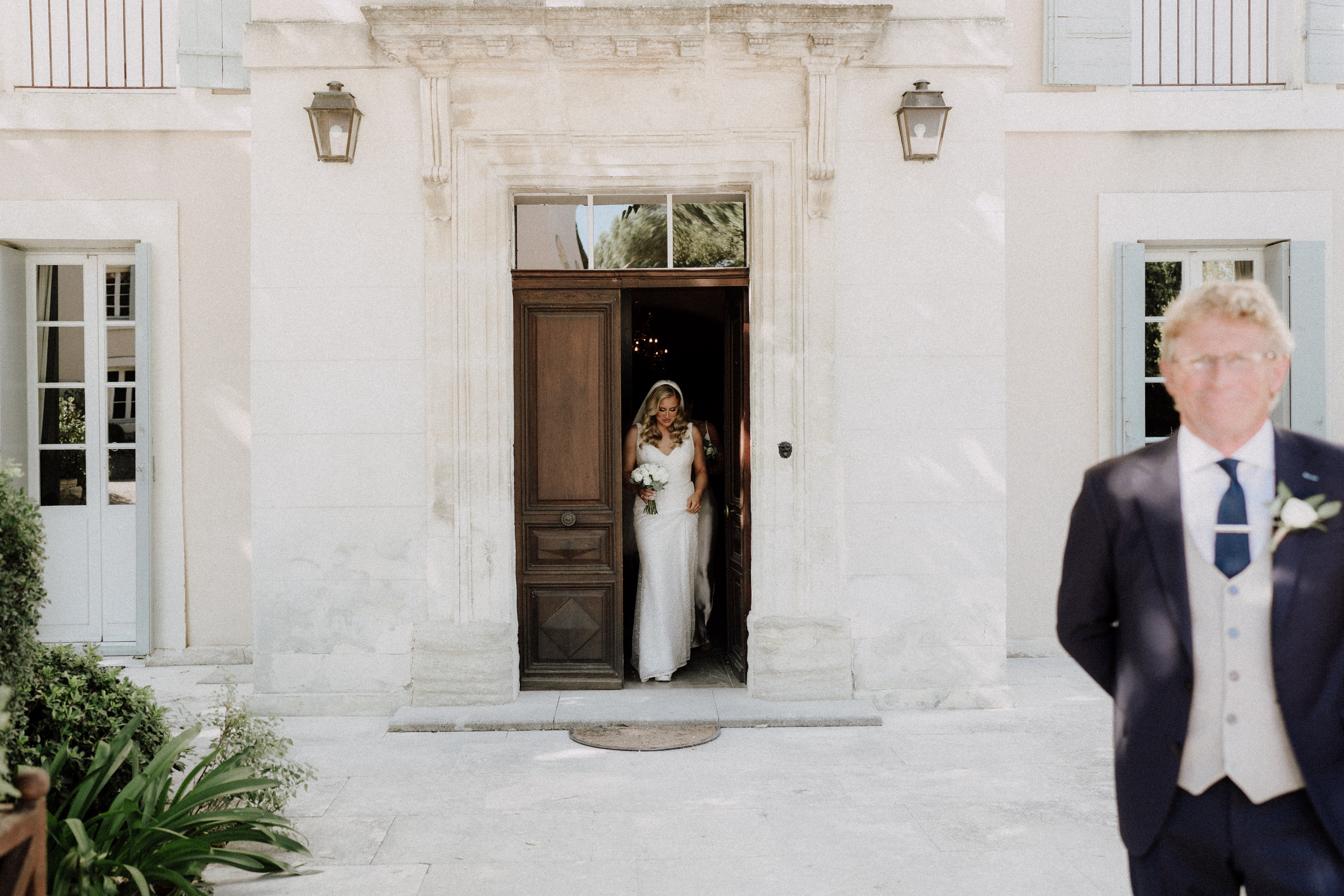 The bride is stepping out through a large wooden double door of a French chateau or manor house, captured in a wide shot that includes the full facade of the building. She is wearing a fitted white lace gown with a V-neckline and a long veil, and is holding a bouquet of white flowers, likely white roses or peonies. In the foreground to the right, an older man in a navy suit with a light grey waistcoat and navy tie — likely the father of the bride — stands with his back partially to the camera, out of focus. The building exterior features pale stone, blue-grey shutters, and two wall-mounted lanterns flanking the ornate doorway. Potential venue feature image.