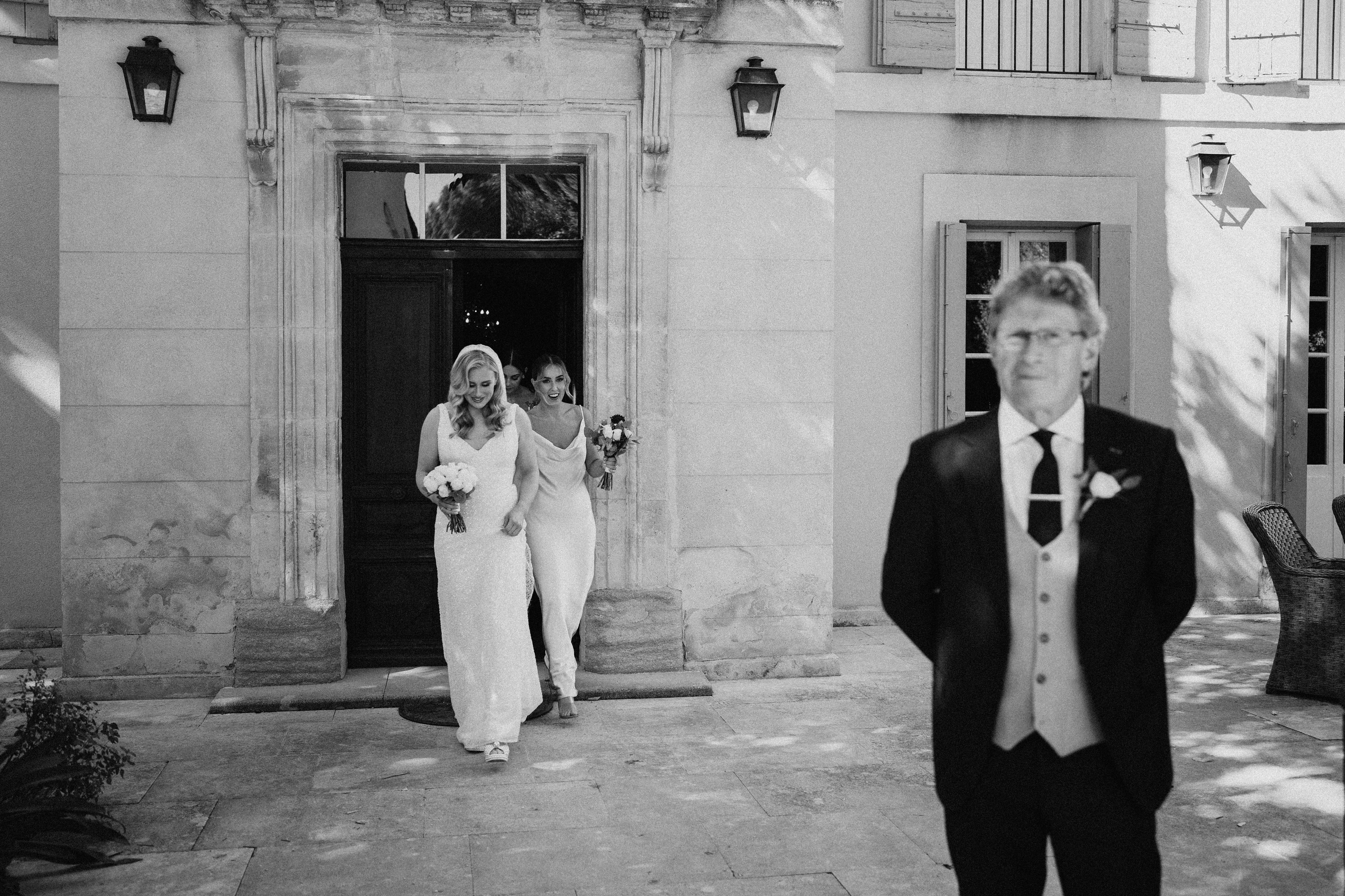 This black-and-white image captures a candid moment of the bridal party emerging through the main entrance of a French chateau or manor house, stepping out onto a stone-paved courtyard. Two women in the foreground wear fitted, floor-length white gowns — one with a veil and lace detailing, the other in a sleek cowl-neck style — both holding bouquets and smiling broadly, with at least one more figure visible in the doorway behind them. In the right foreground, slightly out of focus, stands an older man in a dark suit with a waistcoat, dark tie, and white boutonniere, his back turned to the women. The composition is a wide documentary-style shot with strong contrast between the bright exterior stonework and the dark doorway, framing the bridal party's exit naturally.