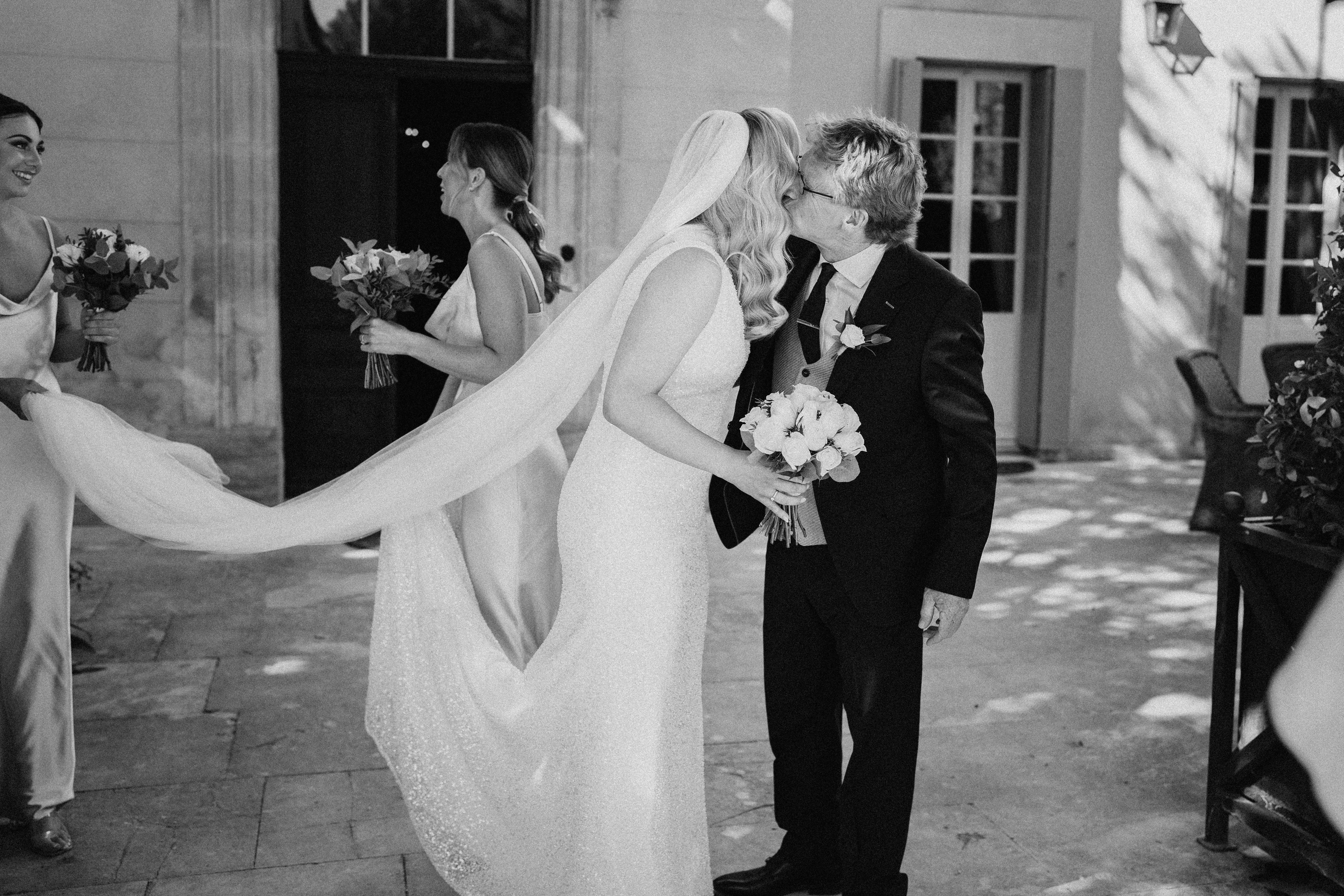 This black-and-white image captures a bride being kissed on the cheek by an older man, likely her father, on an outdoor stone-paved courtyard in front of a classical French chateau or manor building with tall French doors and stone facade. The bride wears a fitted, textured gown with a long flowing veil that is being held aloft by an attendant on the left, and she carries a round bouquet of roses with foliage. The man is dressed in a dark suit with a tie and a boutonniere. Two bridesmaids in satin slip-style gowns are visible in the background and left foreground, each holding smaller bouquets of flowers mixed with greenery. The image is shot in a candid mid-range portrait style with strong contrast between light and shadow, giving it a documentary feel.