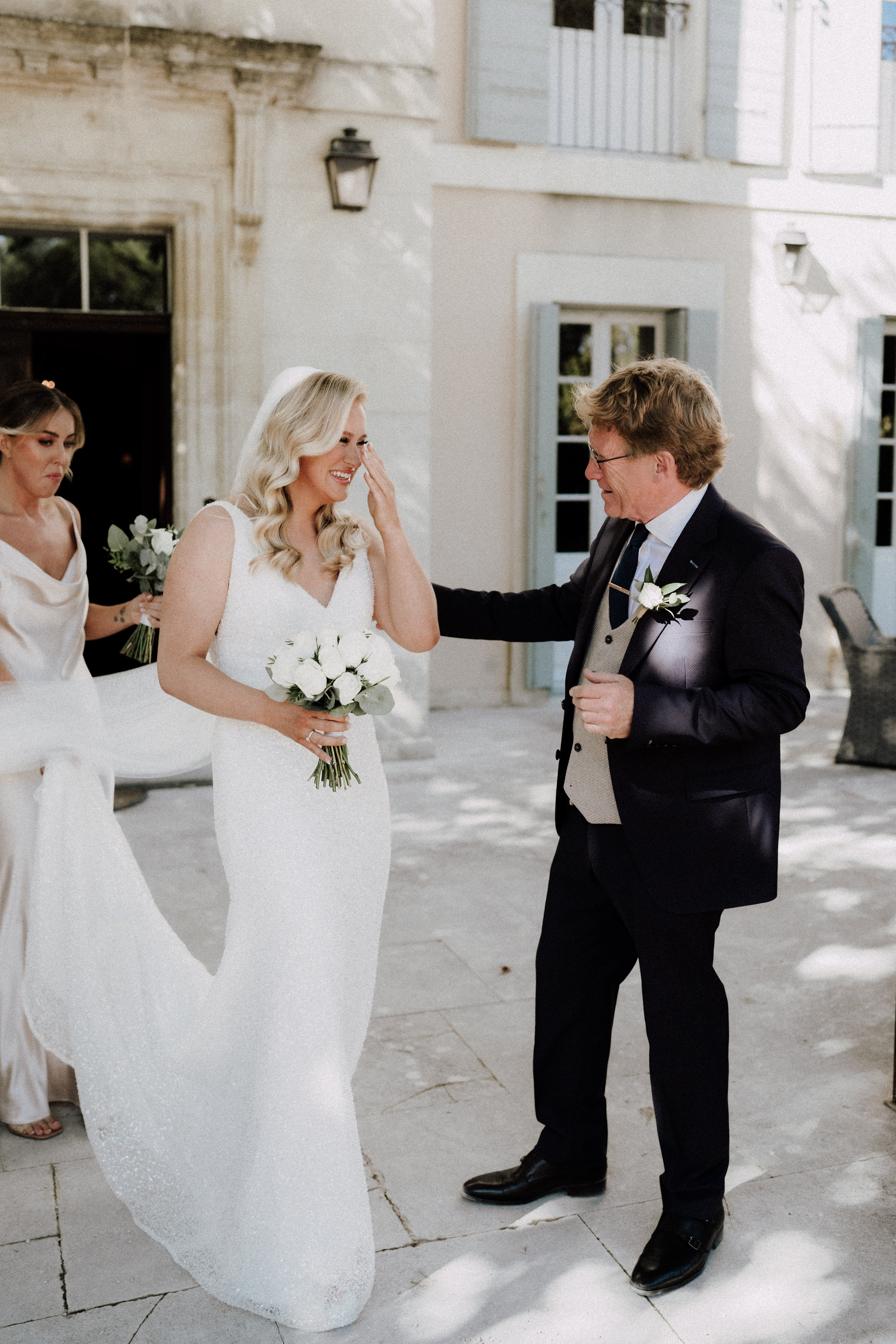 A first look moment between a bride and an older man, likely her father, taking place on the stone terrace of a French chateau with pale blue shuttered windows visible in the background. The bride is wearing a fitted, sleeveless white sequined gown with a V-neckline and holds a compact bouquet of white roses with greenery; she is wiping a tear from her eye while smiling. The man is dressed in a dark navy suit with a beige waistcoat, navy tie, and a white rose boutonniere, and he is reaching out to gently touch her face. A bridesmaid in a champagne/blush cowl-neck dress is partially visible in the background helping with the bride's train. The shot is a candid mid-length portrait captured in natural daylight.
