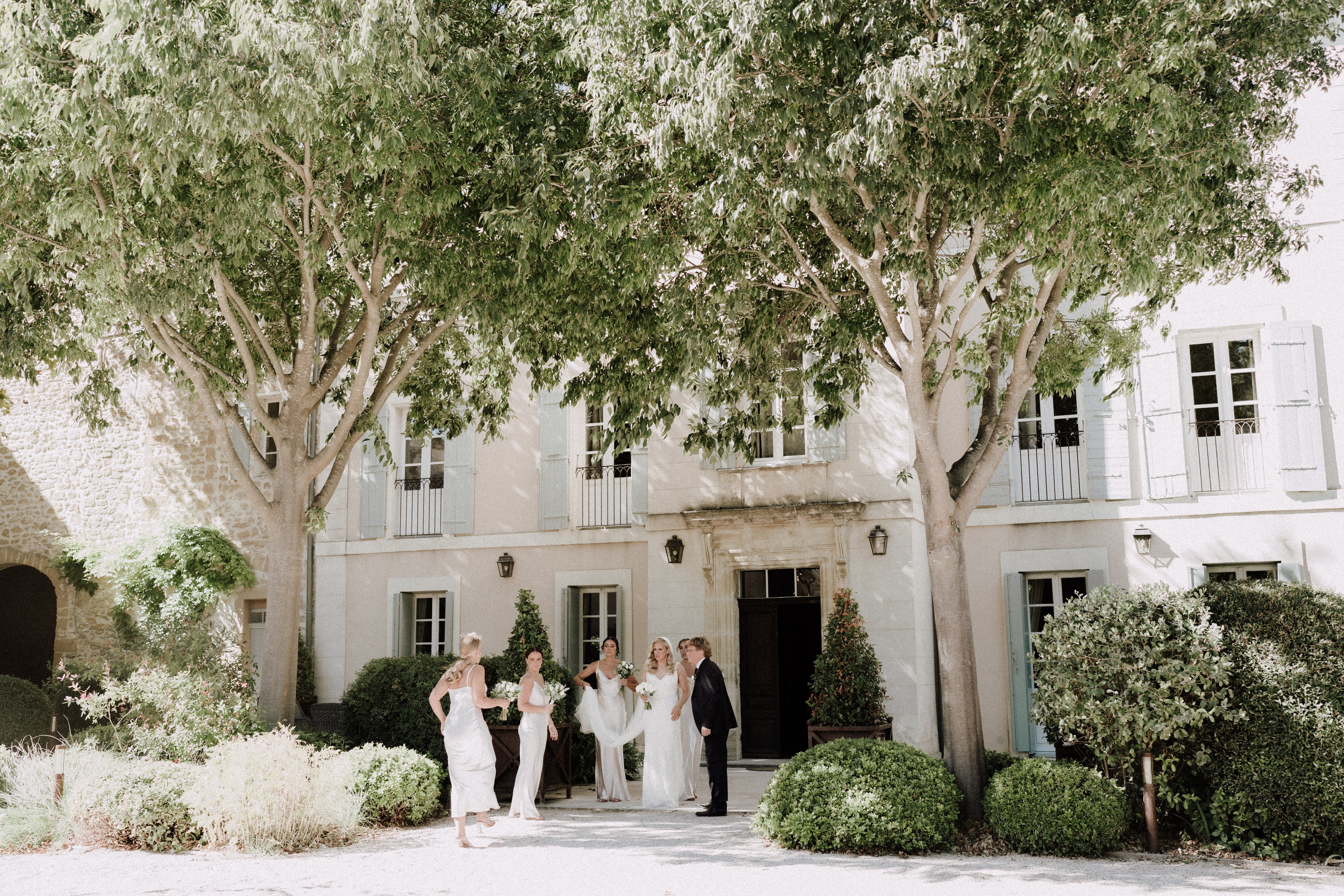 A bridal party of approximately five people gathers outside the main entrance of a French mas or bastide-style property, captured in a wide shot. The bride wears a fitted white gown with a train and carries a bouquet of white flowers with greenery, while three bridesmaids are dressed in ivory or white slip-style dresses; one male guest or escort in a dark suit stands beside the bride. The two-story cream-rendered facade features sage-green shutters, wrought-iron balconies, and classic stone detailing around the entrance doorway, with trimmed boxwood hedges and climbing greenery flanking the door. The overall styling is minimal and classic, with an all-white and ivory palette for the bridal party. Potential venue feature image.